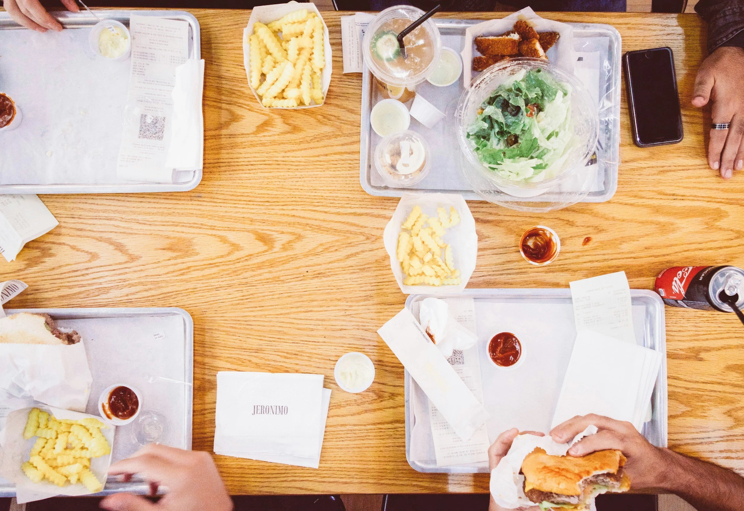 Top-down view of a wooden table with trays of fast food, including fries, salad, burger, soda, and dipping sauces, with people reaching for food and holding a burger, in a casual dining setting.