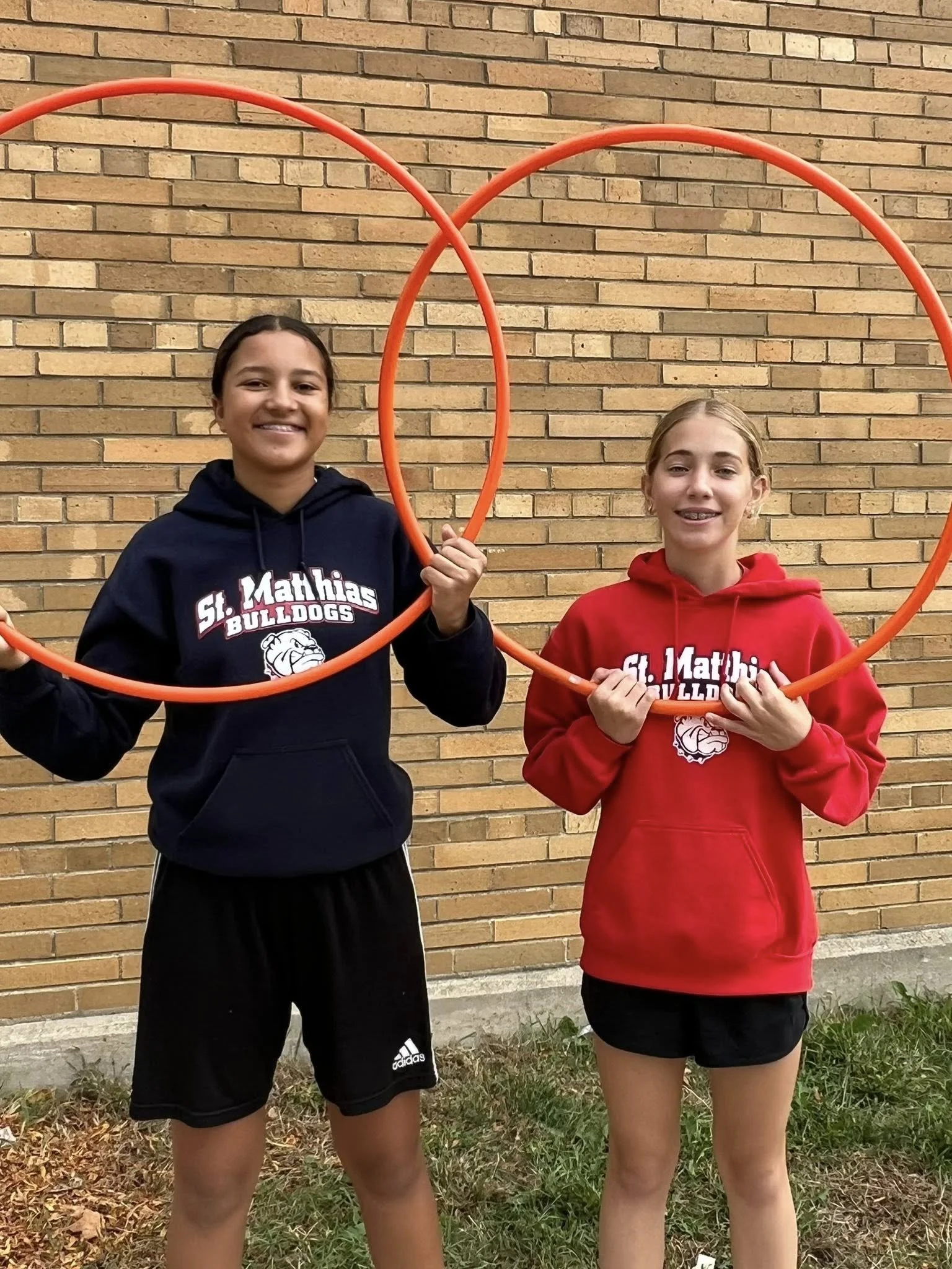 Two young girls standing outdoors in front of a brick wall, holding orange hula hoops. They are smiling and wearing hoodies with 'St. Mathias Bulldogs' logo.
