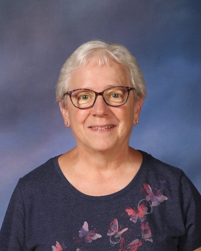 A smiling woman with short gray hair, wearing glasses, earrings, and a navy blue shirt with pink and purple butterfly designs.