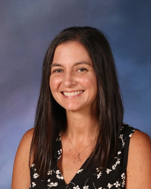 A woman with long dark hair smiling against a blue gradient background, wearing a sleeveless black and white floral top and a gold necklace with a small pendant.