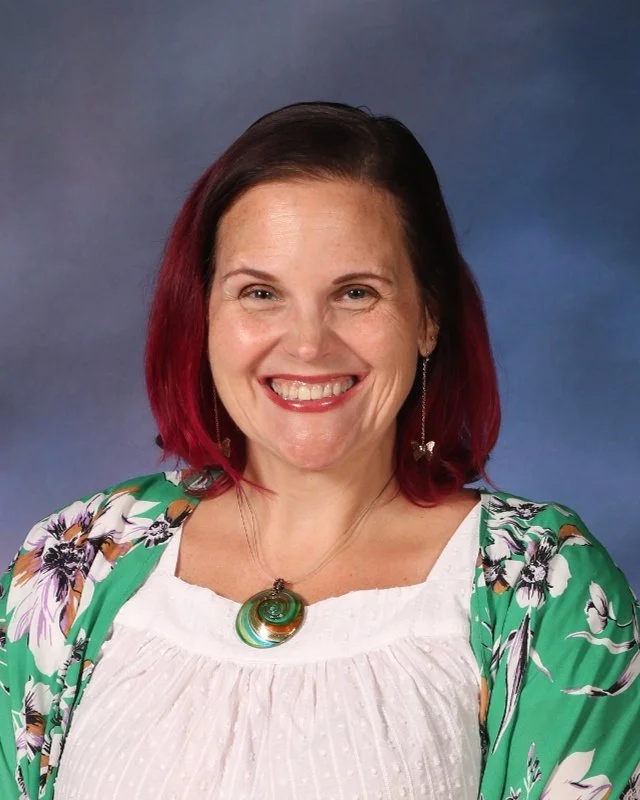 A woman with shoulder-length red hair, smiling, wearing a white blouse, a green floral cardigan, and a large green and gold pendant necklace against a blue gradient background.
