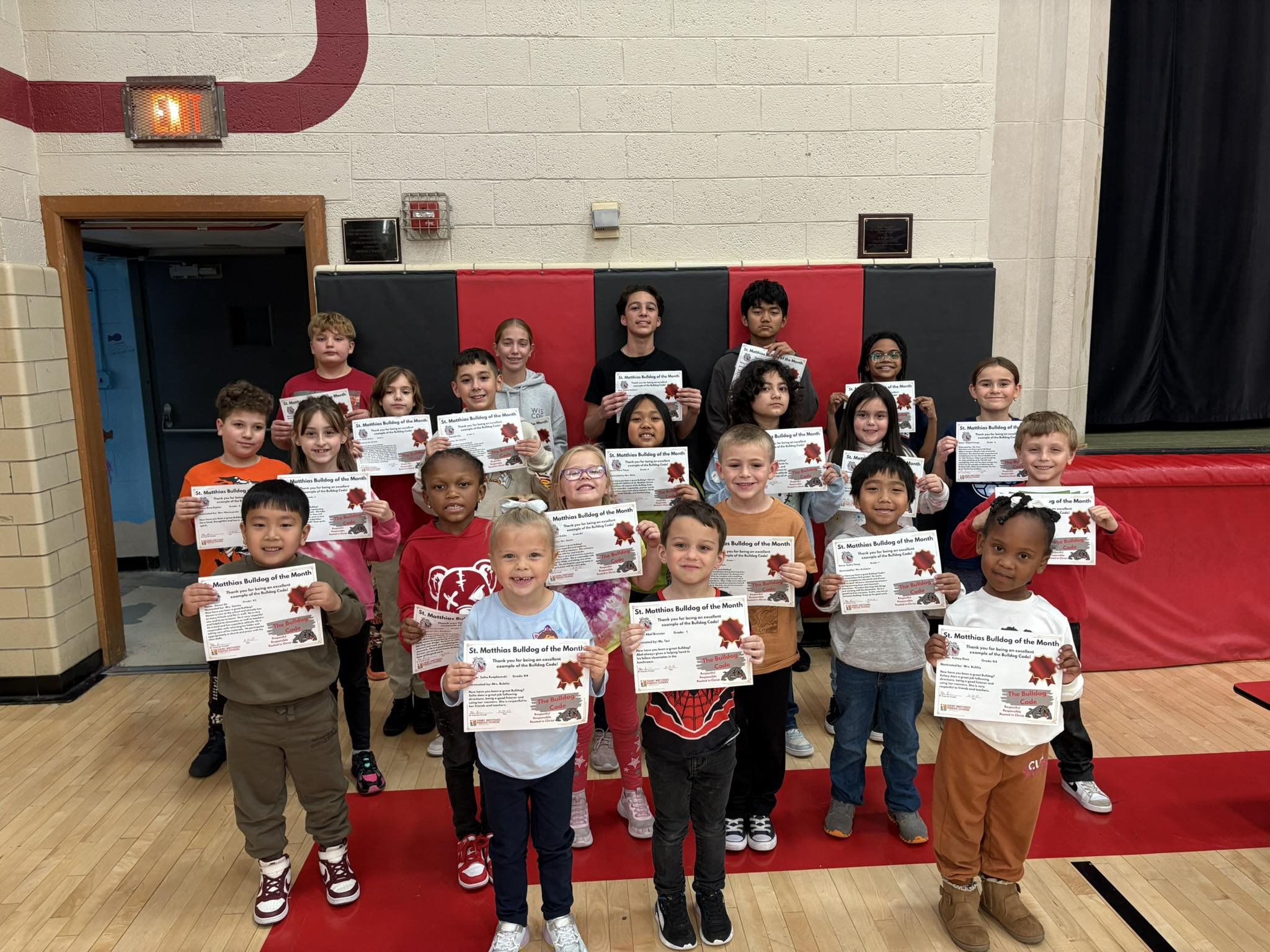 Group of young students standing on a gymnasium floor, holding certificates for accomplishing the goals set in the monthly Bull Dog code, posing for a photo.