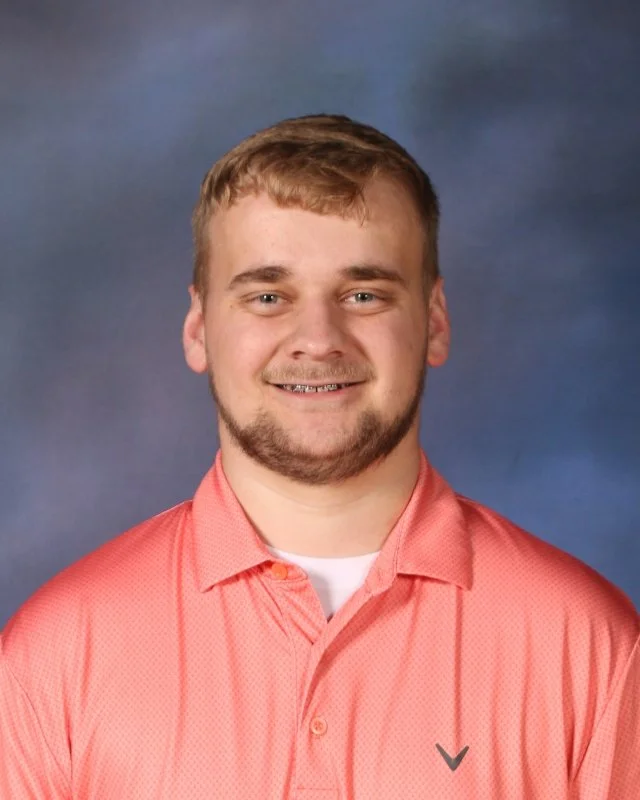 A young man with short, light brown hair and a beard, smiling, wearing a coral button-up shirt with a white T-shirt underneath, standing against a blue background.