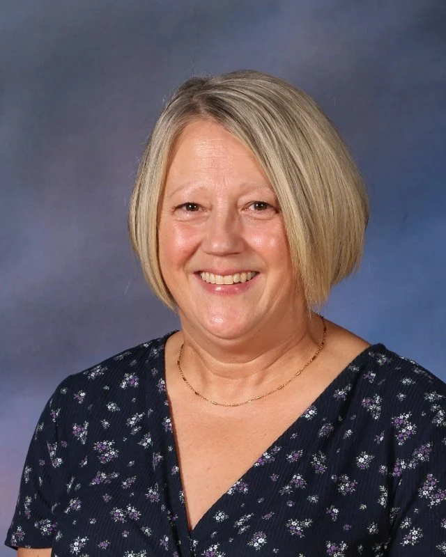 A middle-aged woman with blonde hair styled in a bob, smiling, wearing a black dress with small purple and white floral patterns, and a thin gold necklace against a blue-gray background.