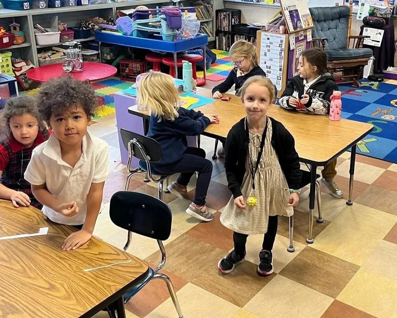 Children in a classroom, some sitting at tables and one girl walking, with bookshelves, toys, and colorful flooring visible in the background.