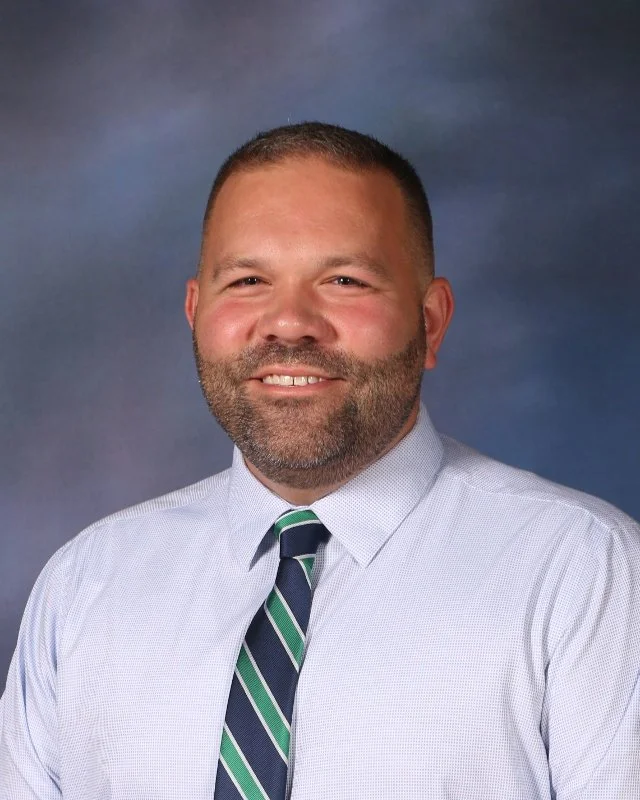A man with short brown hair, a beard, wearing a light blue dress shirt and a green, blue, and white striped tie, smiling against a dark blue background.