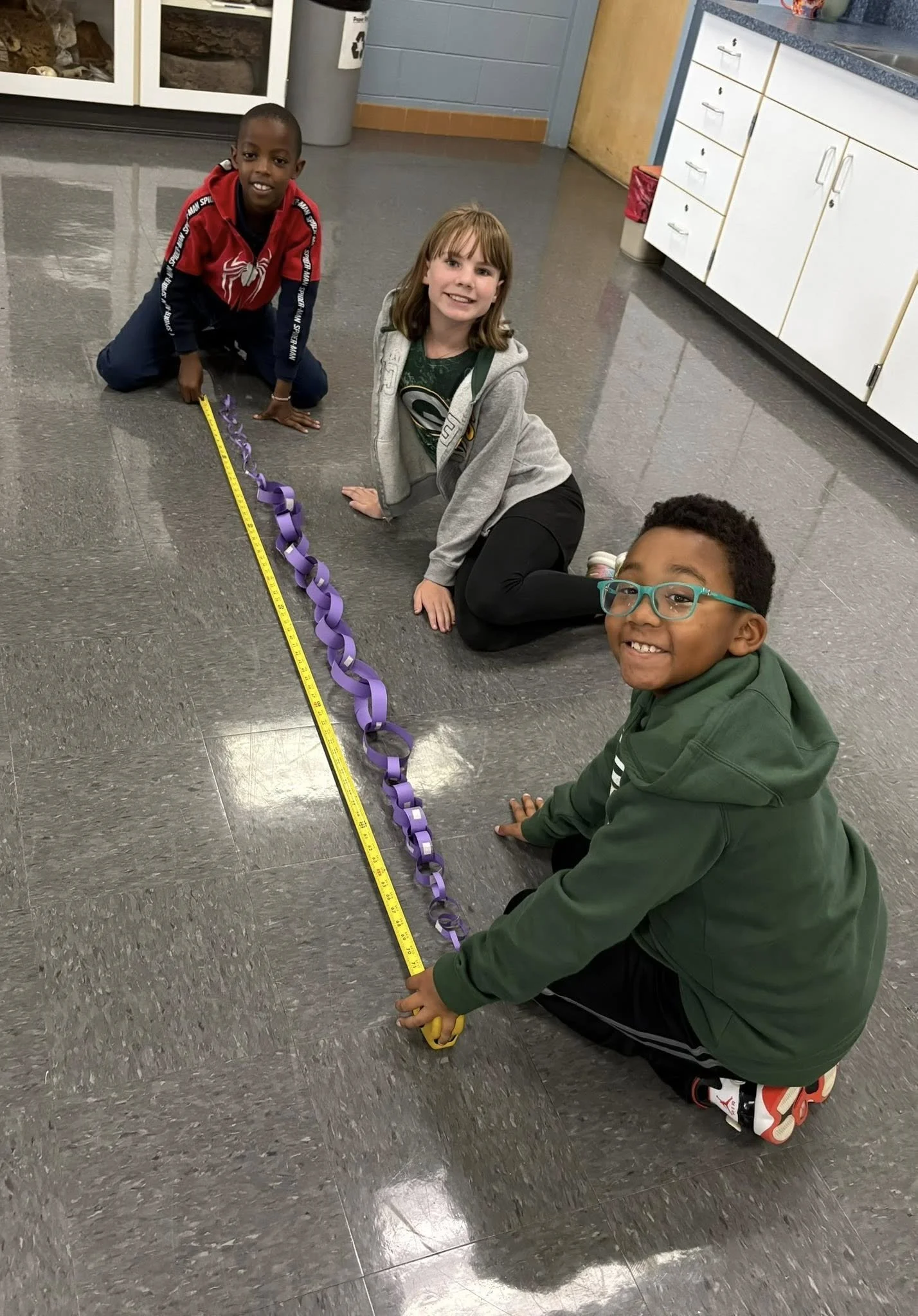 Three children are sitting and kneeling on the floor of a classroom or school setting, with a purple chain and a yellow measuring tape laid out on the floor in front of them, connecting to each other.