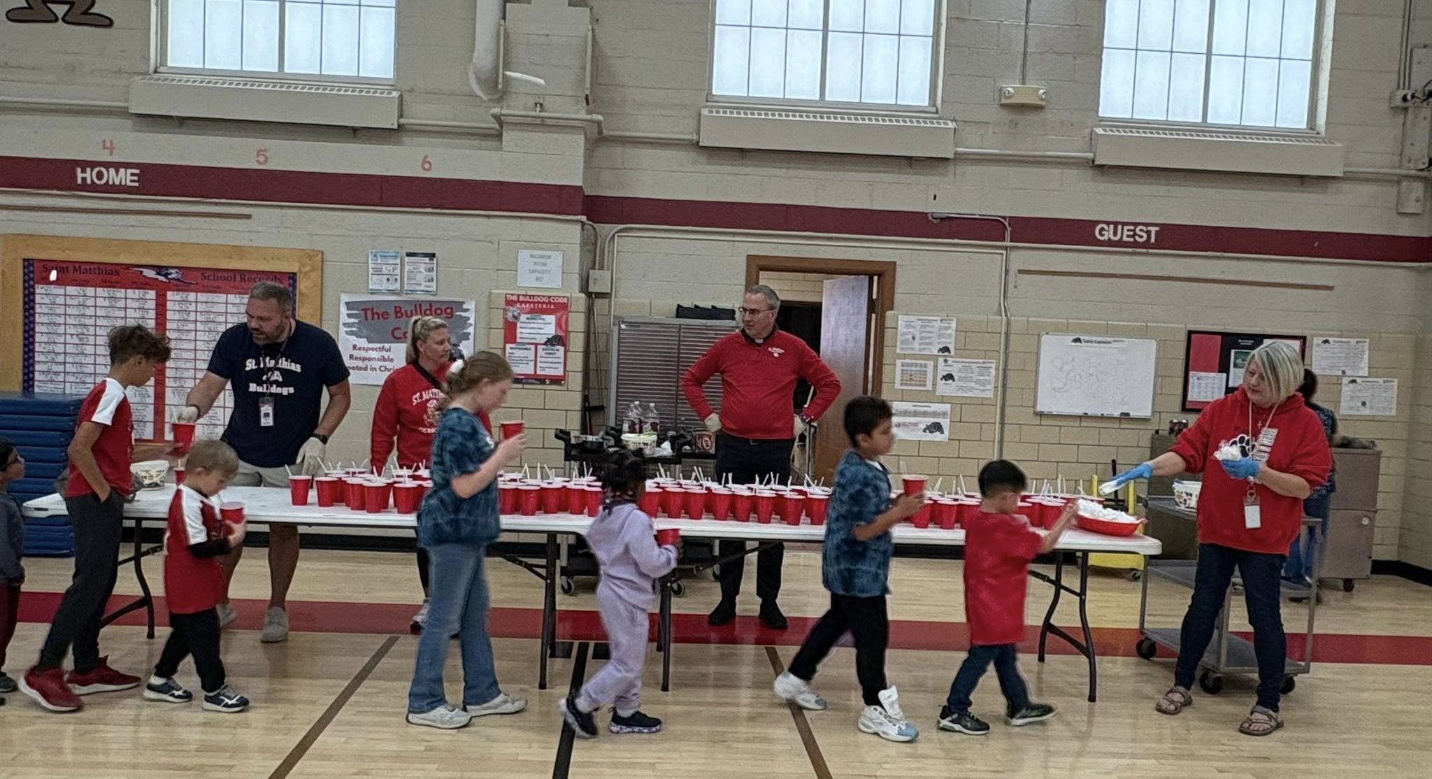 Children lined up in a gymnasium, receiving food served by staff members at a table with red cups. Staff are wearing red and dark blue uniforms, and some are wearing gloves. The background has school notices and signs, including one that says "The Bulldog".