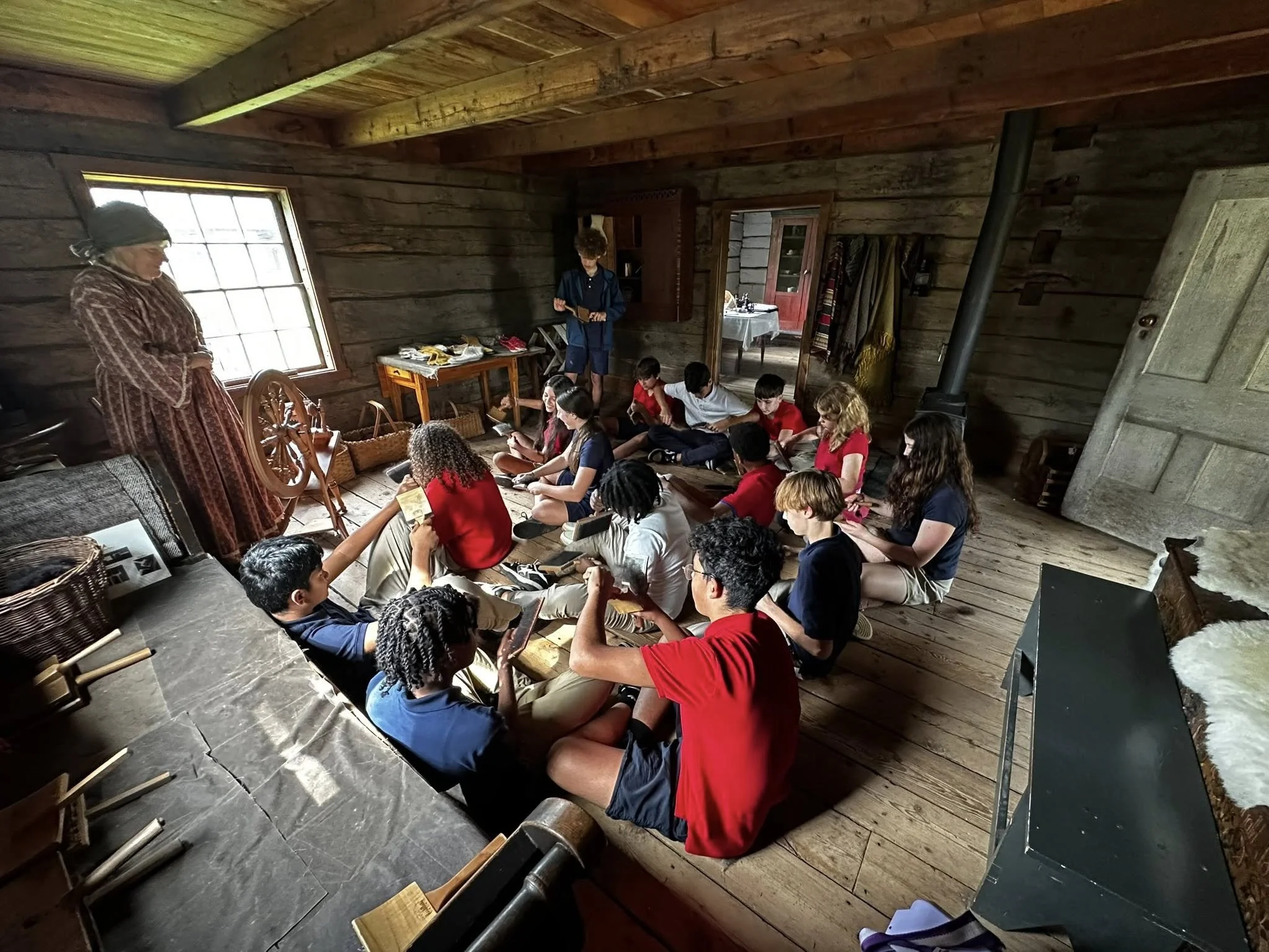 Students visited Old World Wisconsin for a history lesson, they listened to a woman dressed in historical clothing, who appears to be giving a demonstration or storytelling. The cabin has wooden walls, a window, and vintage furnishings.
