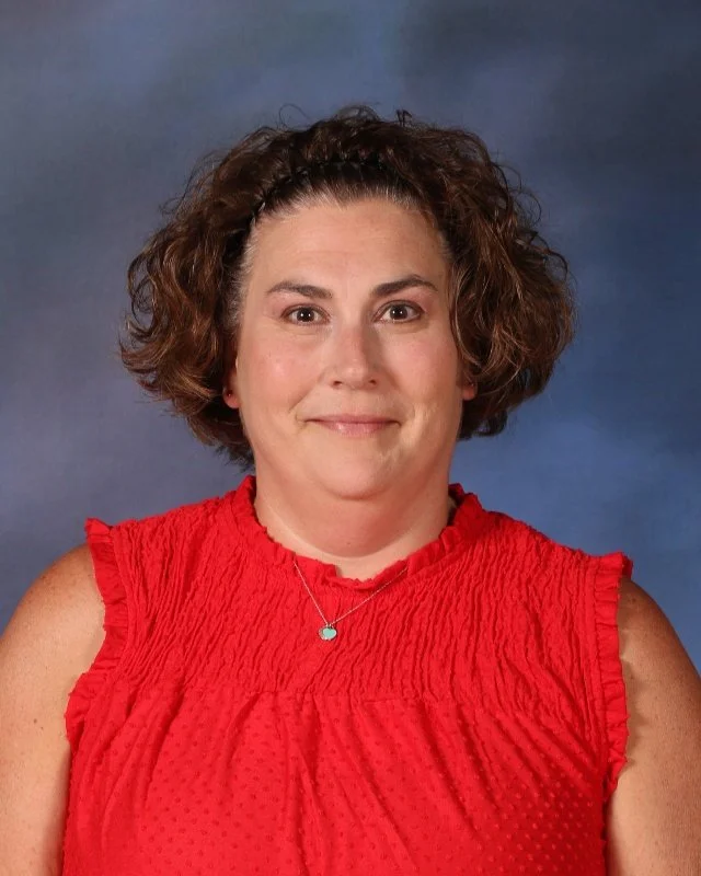 Headshot of a woman with curly brown hair wearing a red sleeveless top and a silver necklace with a small turquoise pendant, against a blue gradient background.