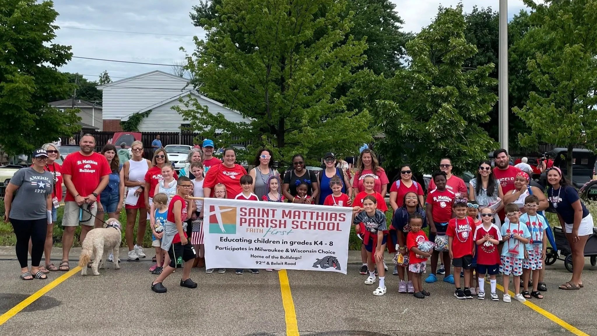 Group of children and adults gathered outdoors holding a banner for Saint Matthias Parish School, with many dressed in red, some wearing sunglasses and backpacks, standing in a parking lot with trees and houses in the background.