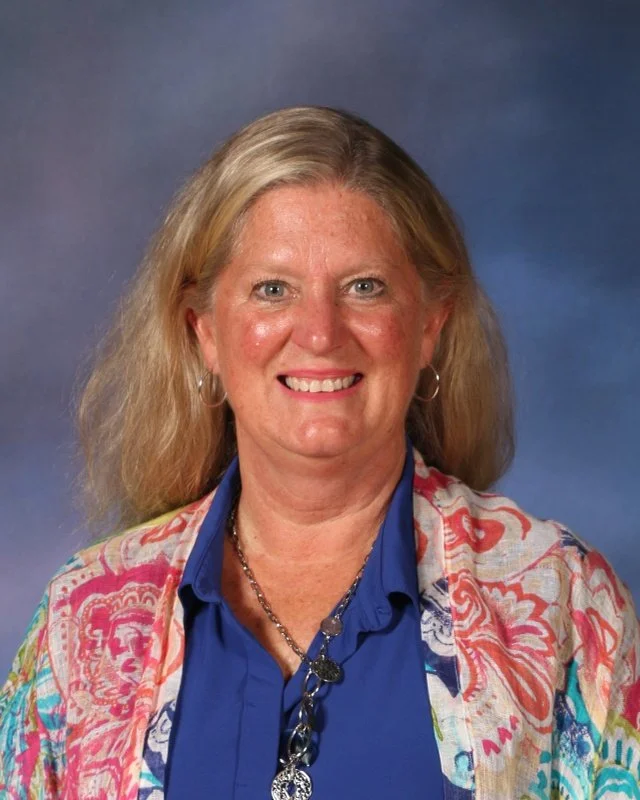 A middle-aged woman with shoulder-length blonde hair, blue eyes, and a warm smile, wearing a vibrant, multicolored patterned blazer over a bright blue blouse, accessorized with silver hoop earrings and a silver necklace, standing against a blue gradient background.
