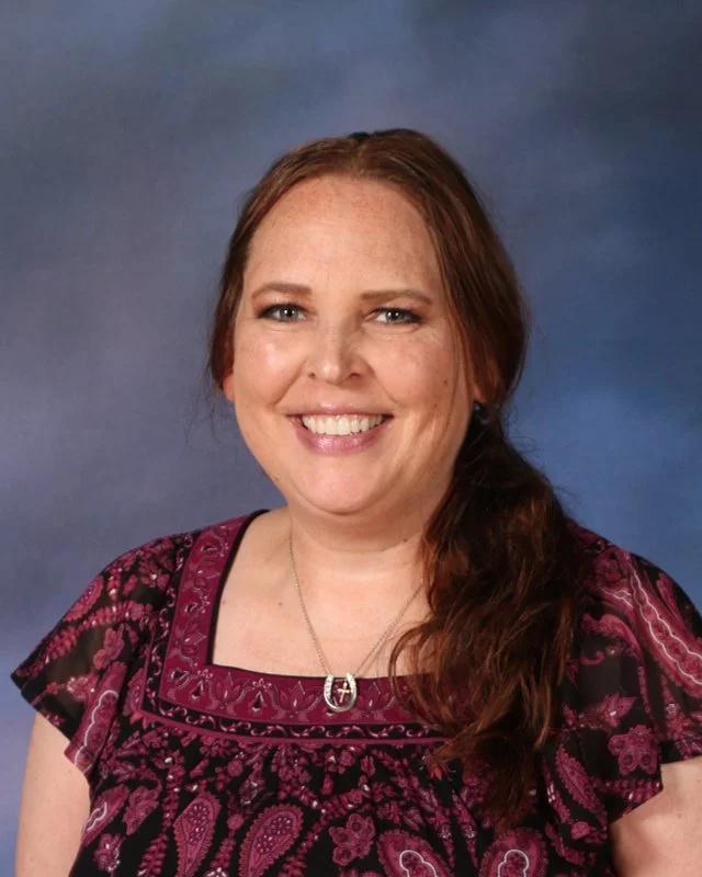 A woman with long brown hair smiling, wearing a black and pink patterned blouse and a silver necklace, against a blue gradient background.