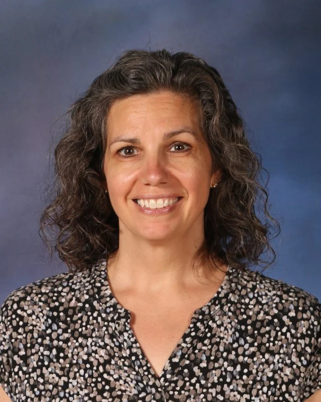 A woman with shoulder-length curly hair smiling, wearing a black and white polka dot blouse, against a dark blue background.
