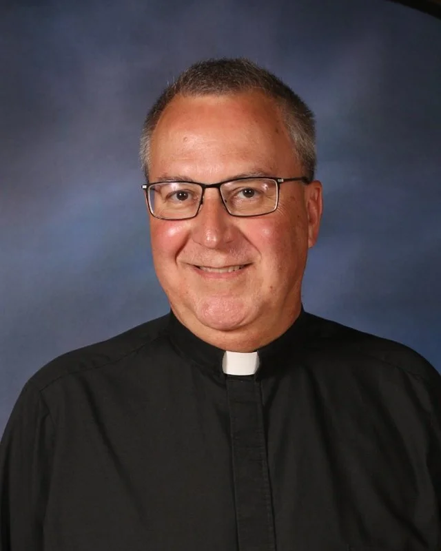 A middle-aged man in clerical attire smiling with gray hair and glasses against a blue background.