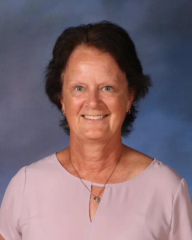 A middle-aged woman with short brown hair, blue eyes, wearing a light purple blouse and small earrings, smiling in front of a blue gradient background.