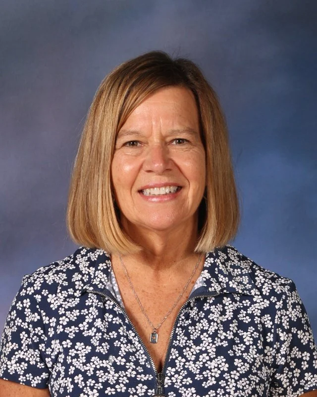 A woman with shoulder-length blonde hair wearing a navy blue floral zip-up shirt and a necklace, smiling against a gradient blue background.