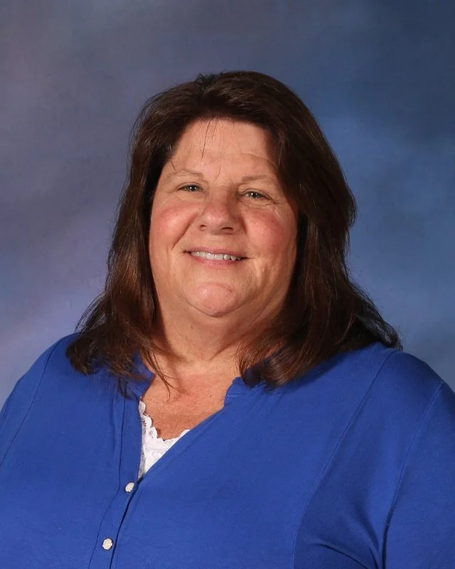 Portrait of a middle-aged woman with shoulder-length brown hair smiling, wearing a blue blouse against a blue gradient background.