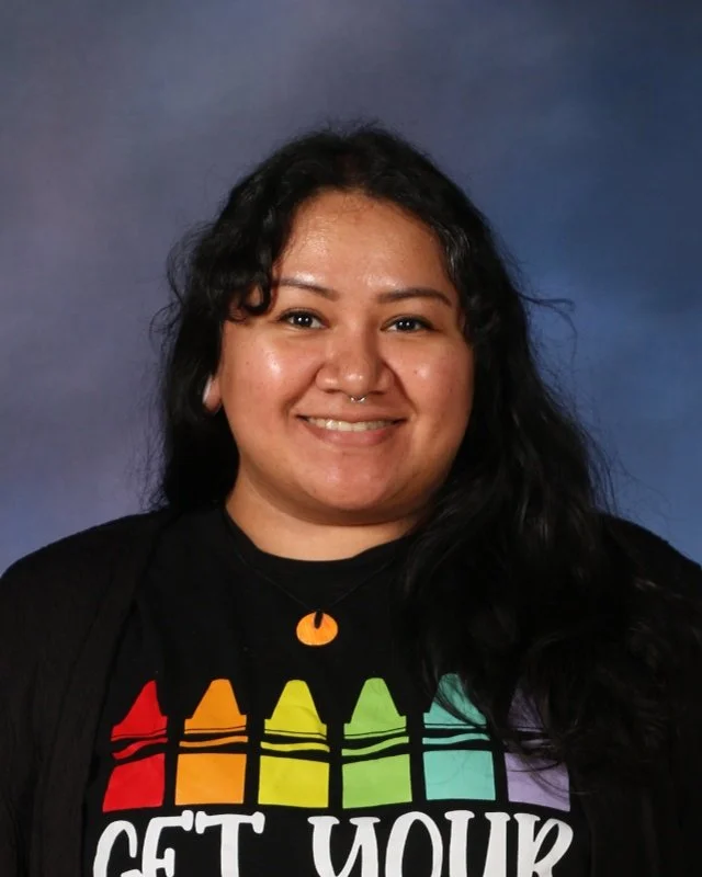 A woman with long black hair, smiling, wearing a black shirt with a rainbow-colored crayon design and the phrase 'Get Your' visible, standing against a blue background.