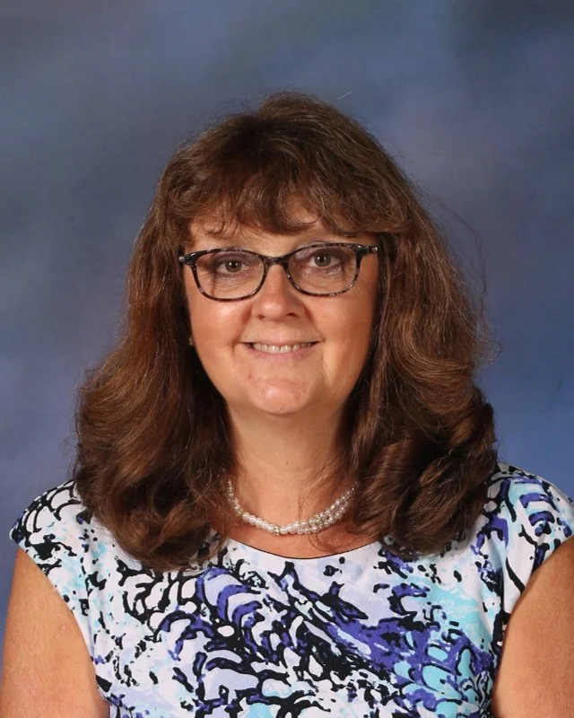 A woman with shoulder-length brown hair, glasses, and a pearl necklace, smiling at the camera, wearing a patterned blouse with blue, black, and white colors, against a blue gradient background.