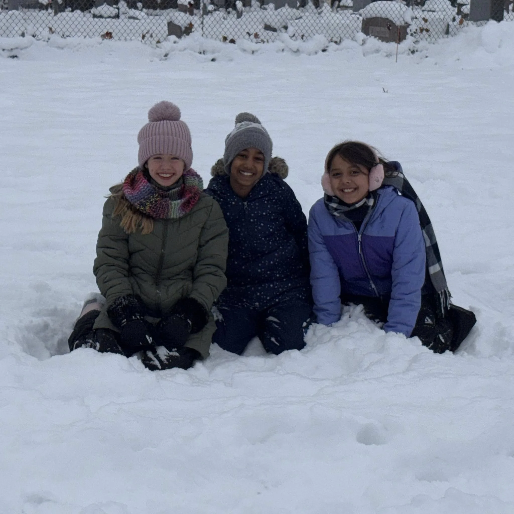 Playing in the snow at recess time. Three girls kneeling in snow, smiling, dressed warmly with hats, gloves, and jackets, in a snowy outdoor setting.