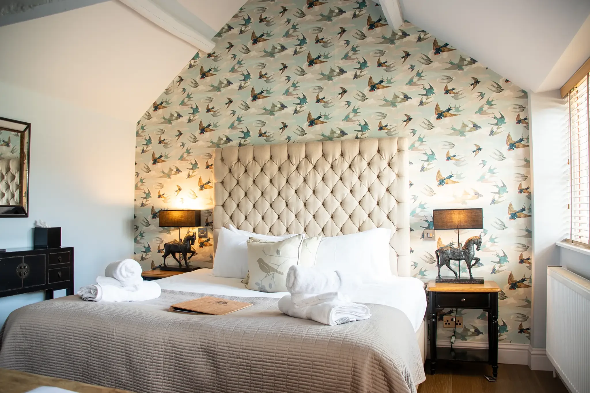 A bedroom with a large, tufted beige headboard, white bedding, and towels shaped like swans on the bed. The wall behind the bed is decorated with bird-themed wallpaper, and there are two nightstands with horse lamp decorations.