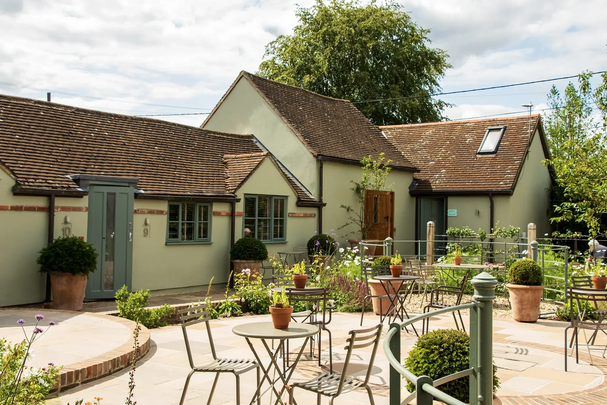 Outdoor patio with metal tables and chairs, potted plants, and a yard with green shrubs, flowers, and a house with a green facade and brown tiled roof under partly cloudy sky.