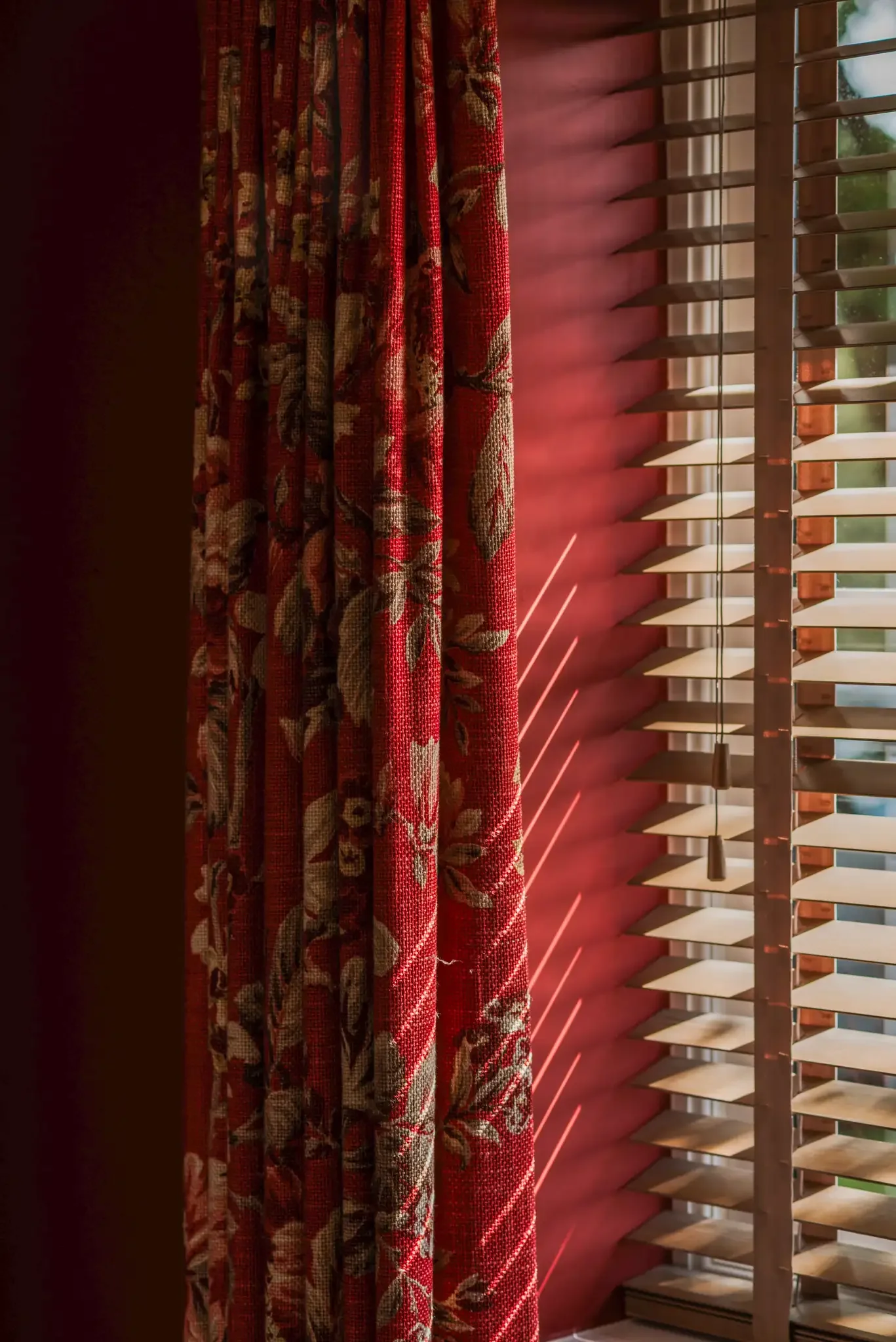Sunlight streams through wooden blinds, casting light and shadows on a red floral curtain and a red wall.