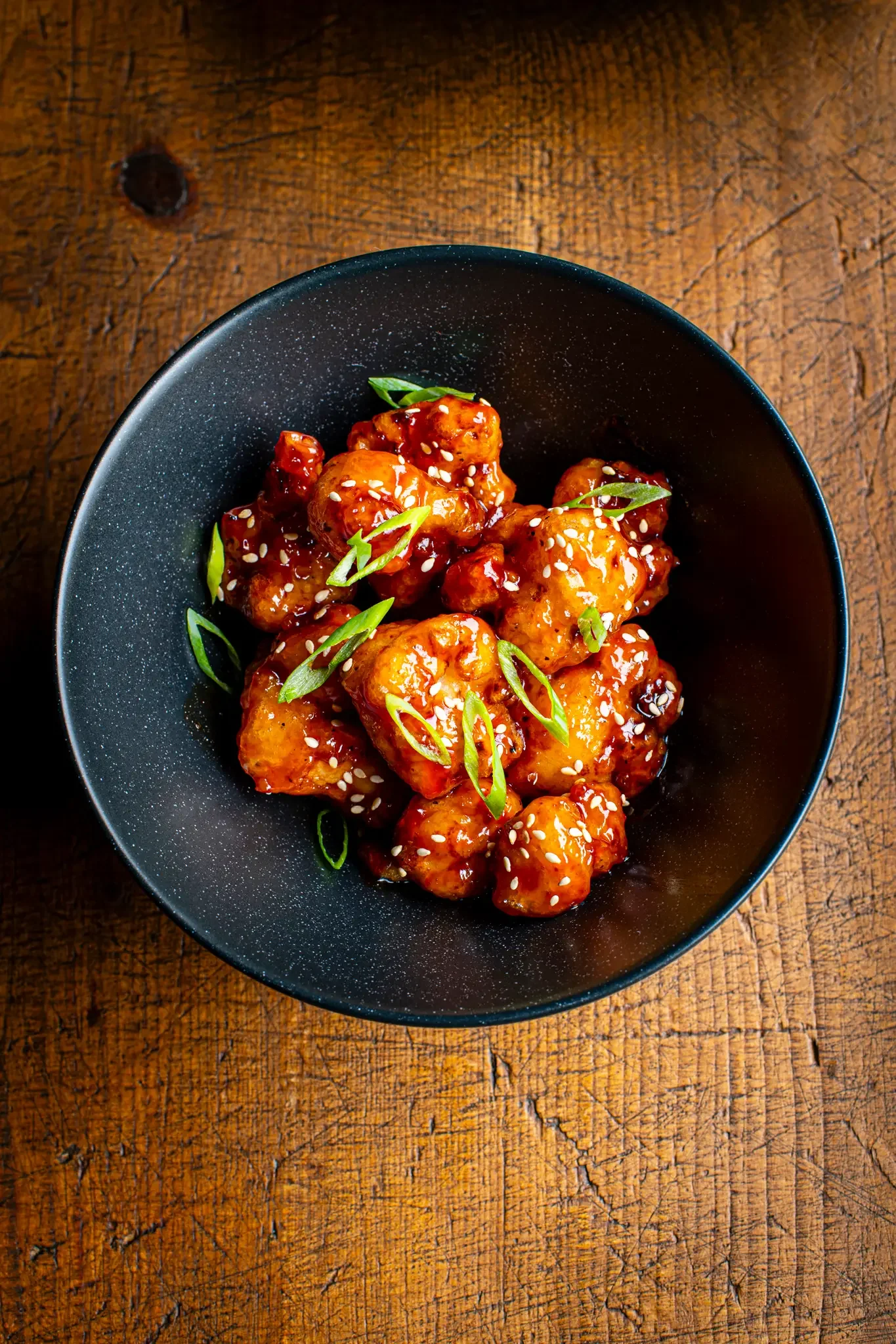 A black bowl filled with sweet and sour chicken garnished with sliced green onions and sesame seeds, placed on a wooden table.