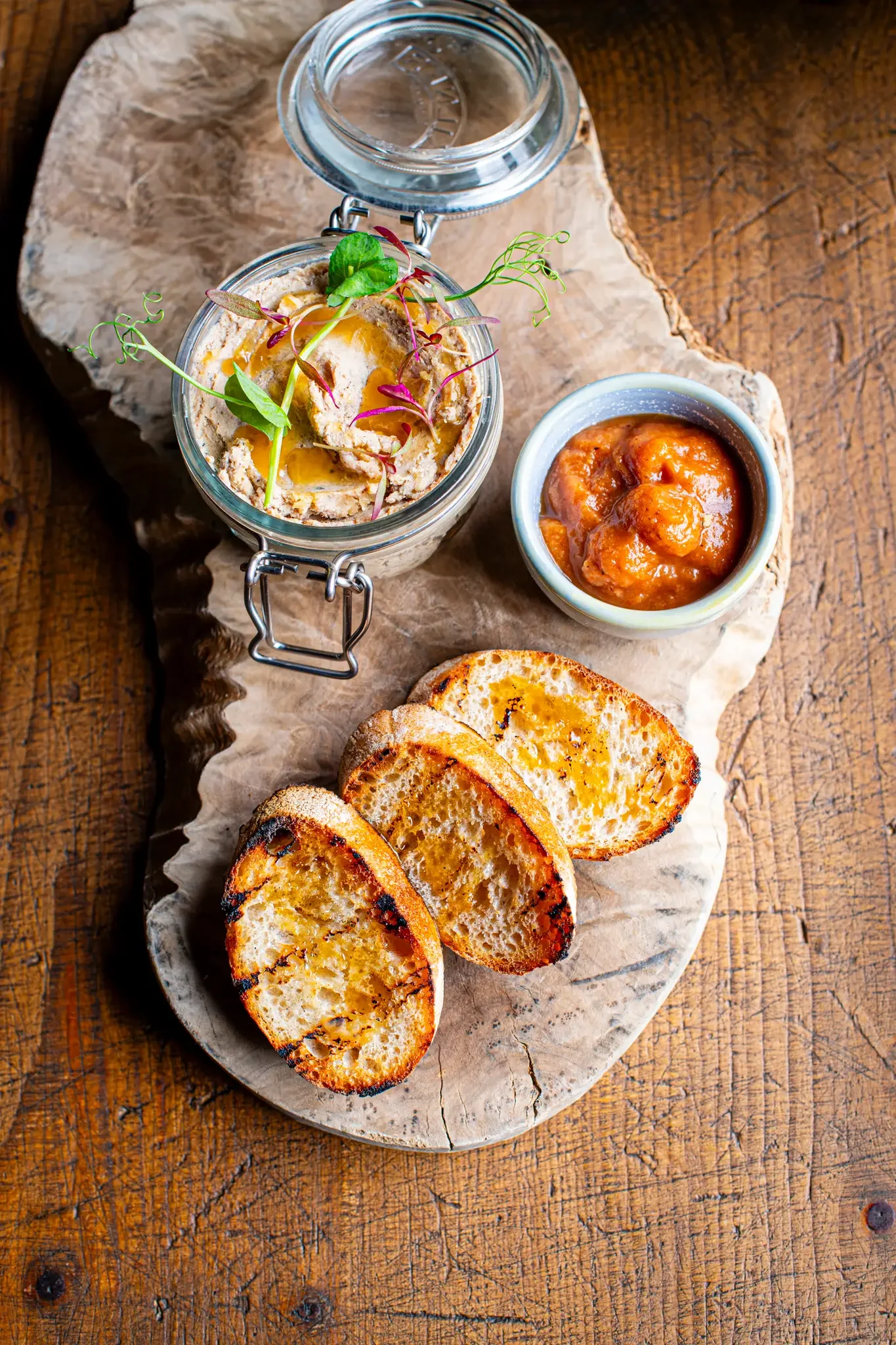 Slices of toasted bread with oil, a jar of hummus topped with herbs and drizzled with oil, a small bowl of chickpea stew or curry, and an empty glass jar on a wooden serving board.