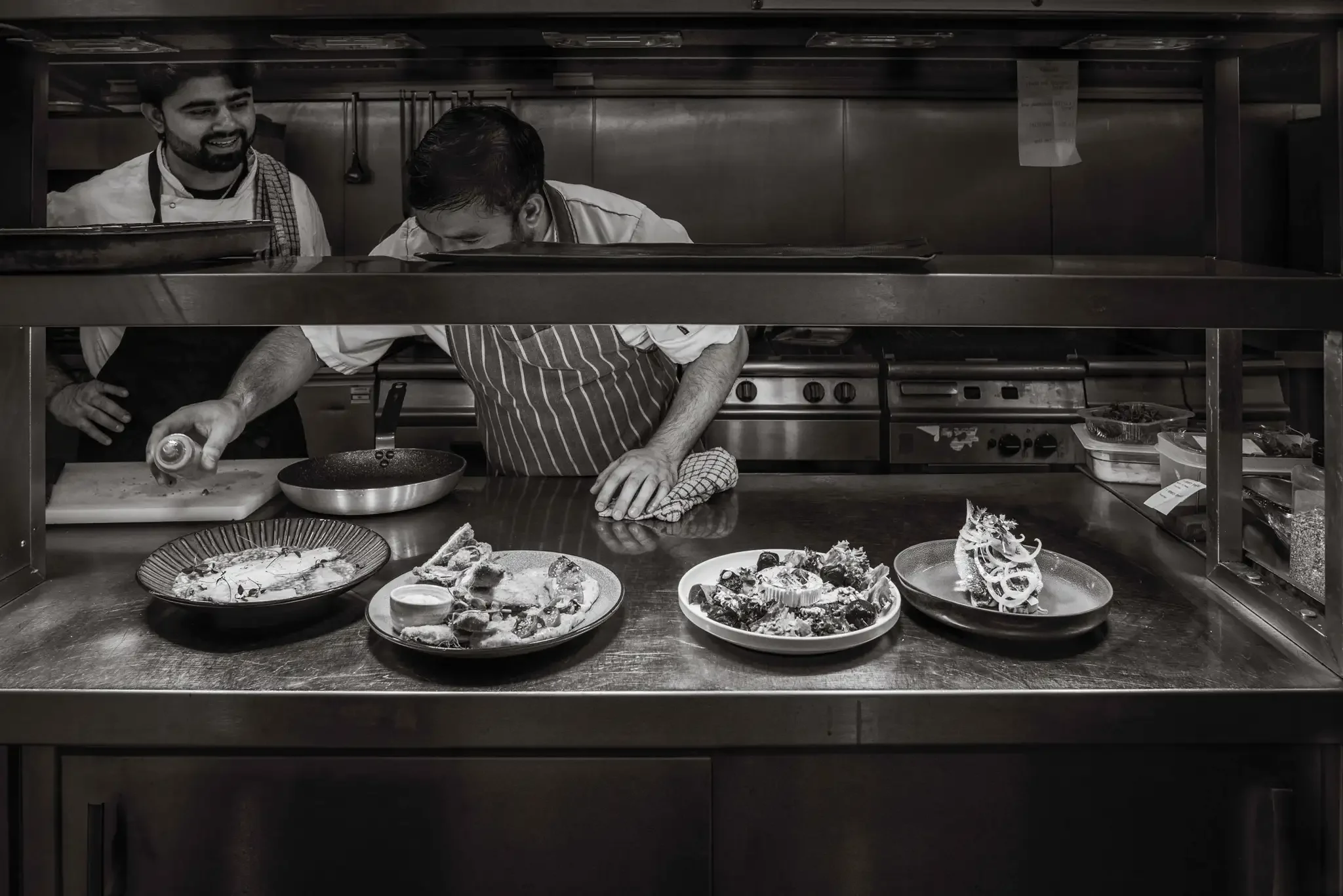 Chefs preparing and garnishing dishes in a professional kitchen, with plates of food on the counter in front of them.