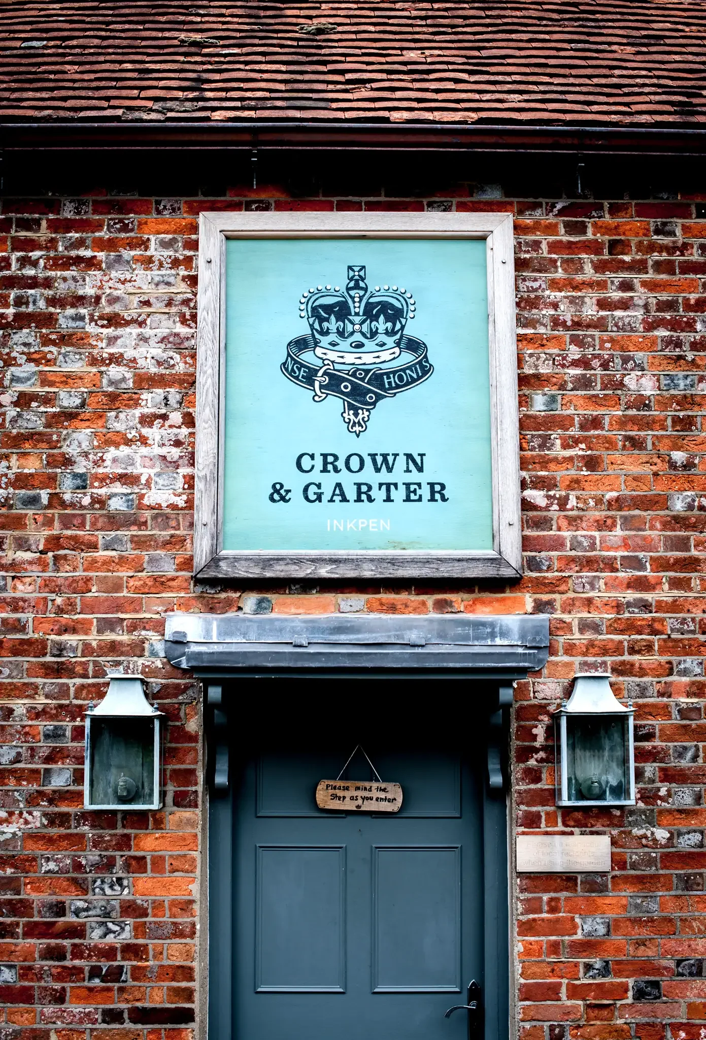 Brick building with a blue sign featuring a crown and sword, reading 'Crown & Garter INKPEN'; dark blue door underneath with a small wooden sign.