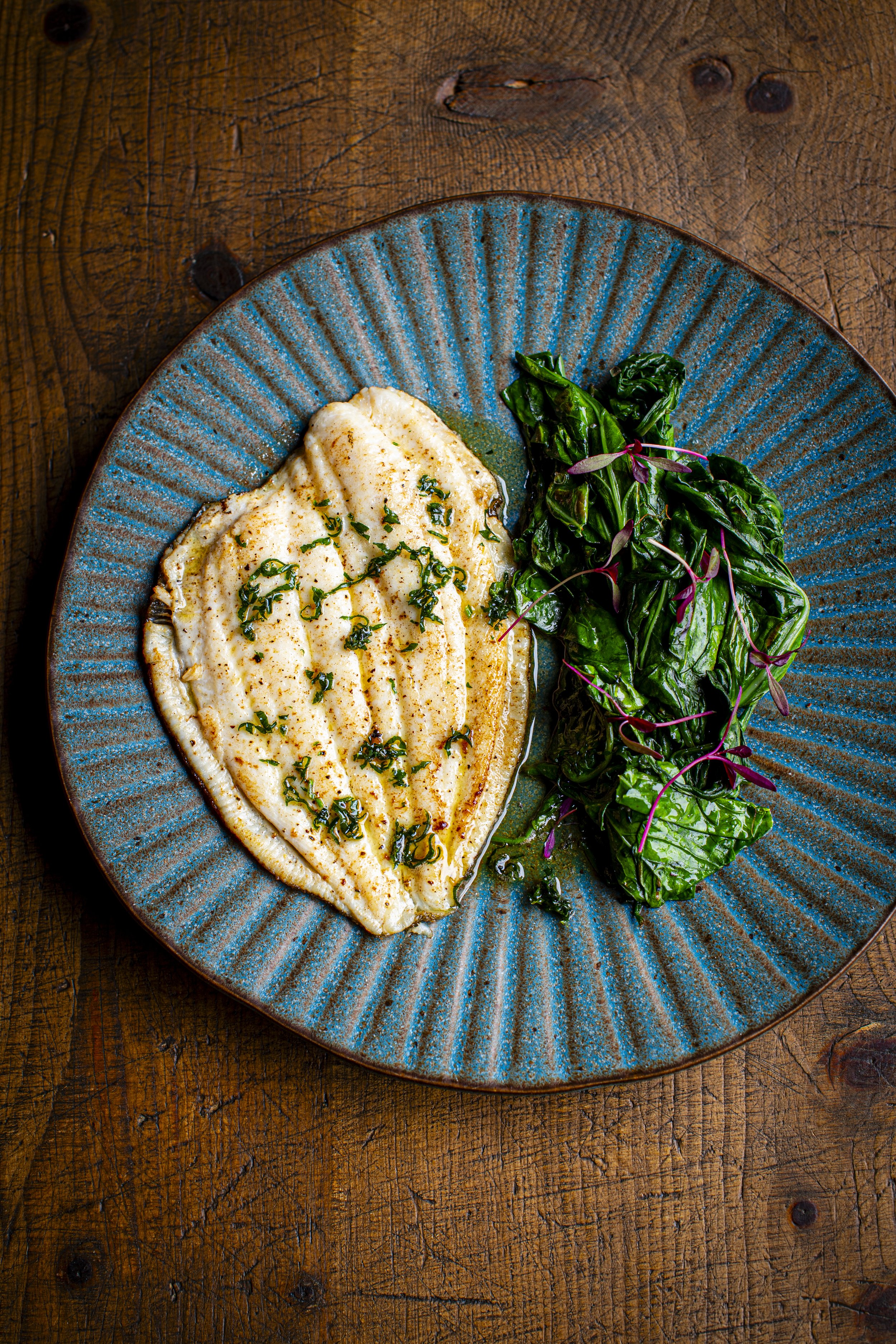 A plate with grilled fish fillet garnished with herbs and a side of leafy greens served on a textured blue plate, placed on a wooden table.