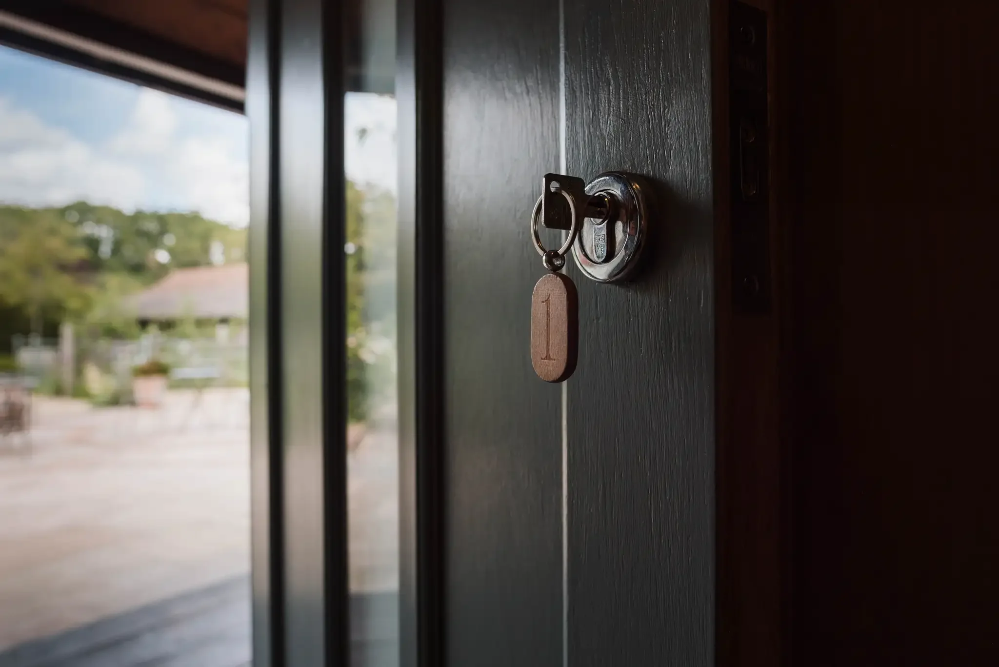 Close-up of a black door with a silver lock and a wooden tag with the number 1 hanging from it, with a blurry outdoor scene visible through a glass panel.