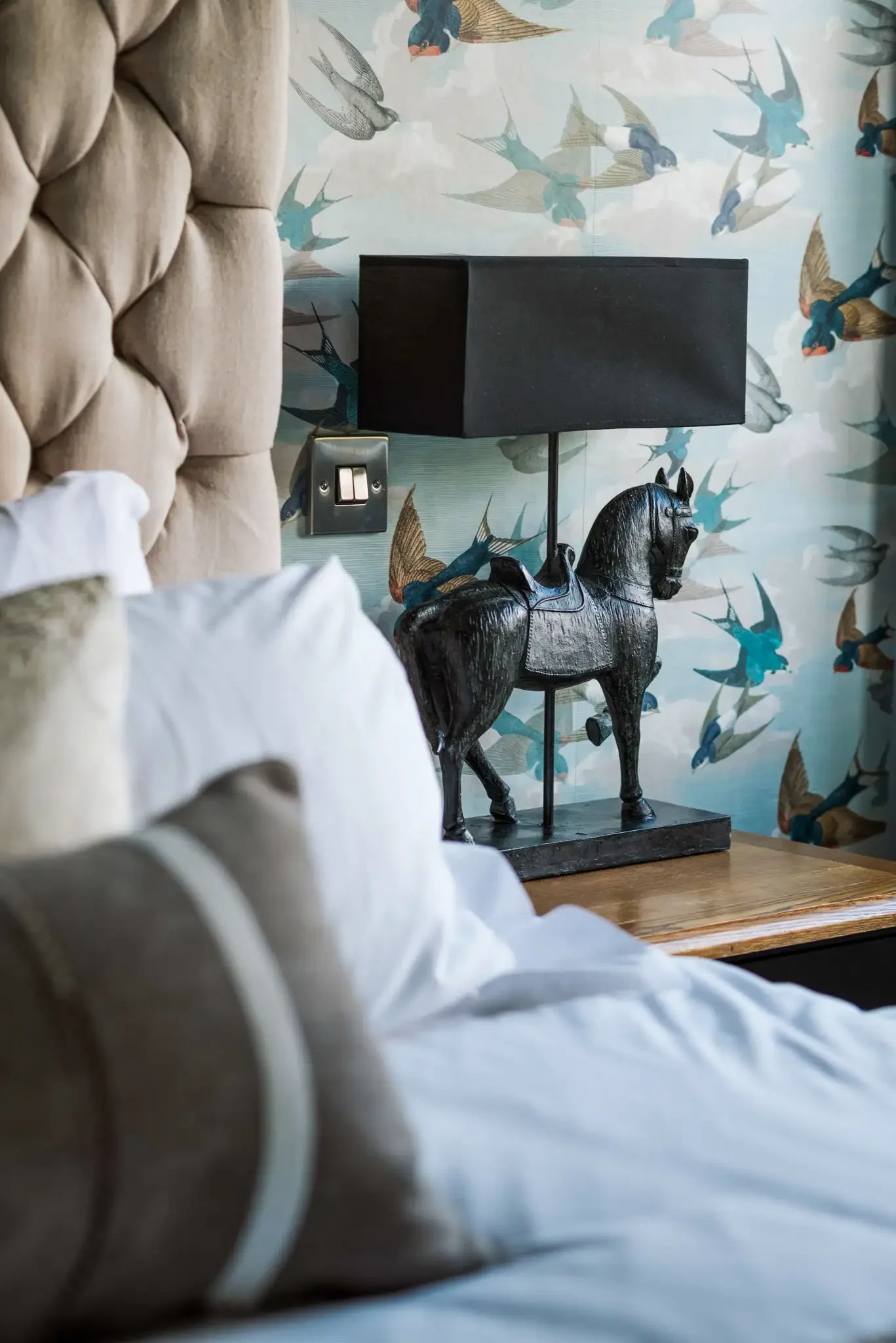 A bedroom with a beige tufted headboard, white bedding, and decorative pillows. There is a wooden bedside table with a black horse-shaped lamp and a black lampshade. The wall behind has wallpaper with a bird pattern.