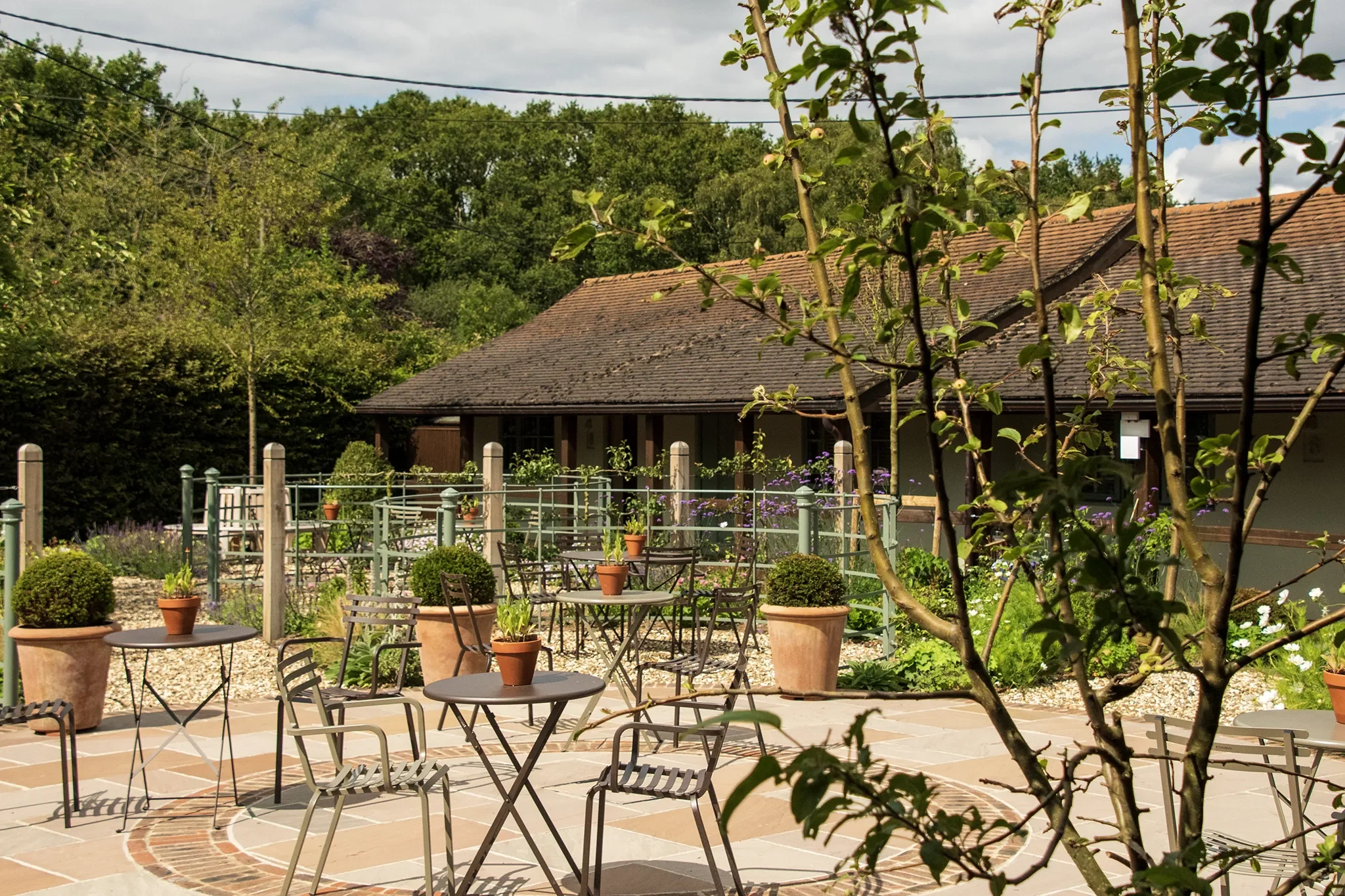 Outdoor patio area with metal tables and chairs, potted plants, trees, and a building with a tiled roof in the background under a cloudy sky.