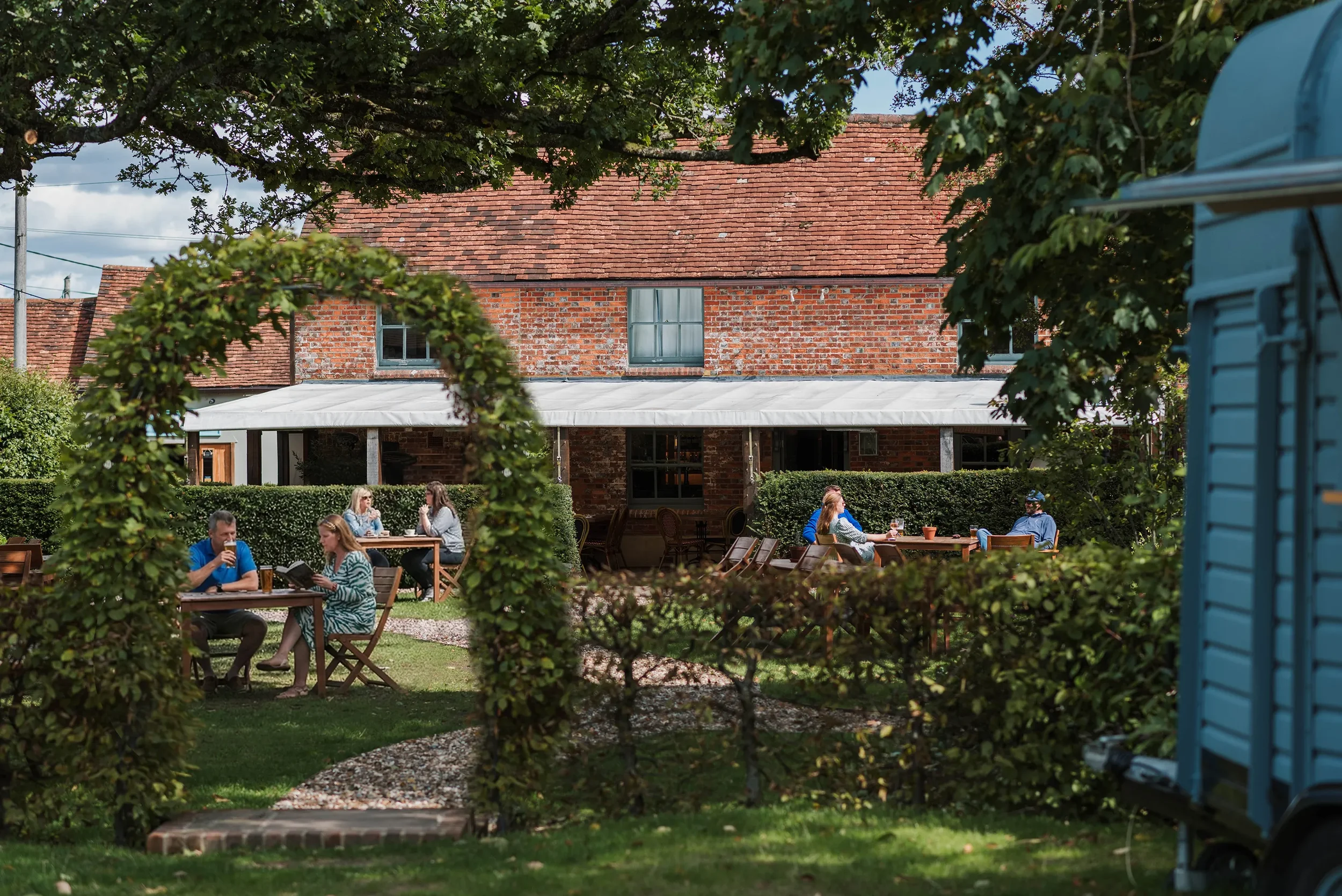 People dining outdoors in a garden patio with a brick house in the background, framed by green leafy bushes and a wooden archway, on a partly cloudy day.