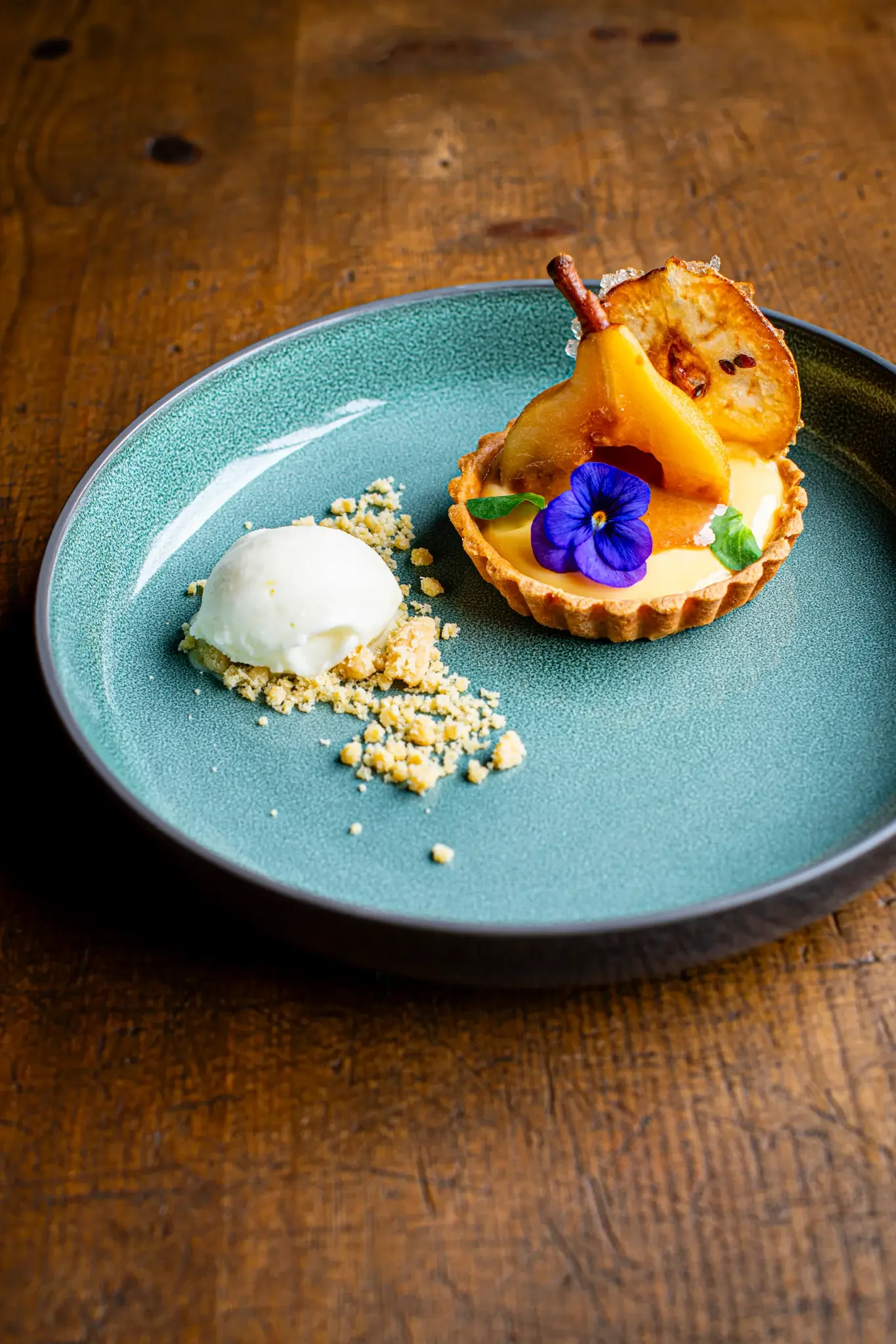 A dessert tart with a pear, garnished with an edible flower and mint leaves, served with a scoop of vanilla ice cream on a teal plate.