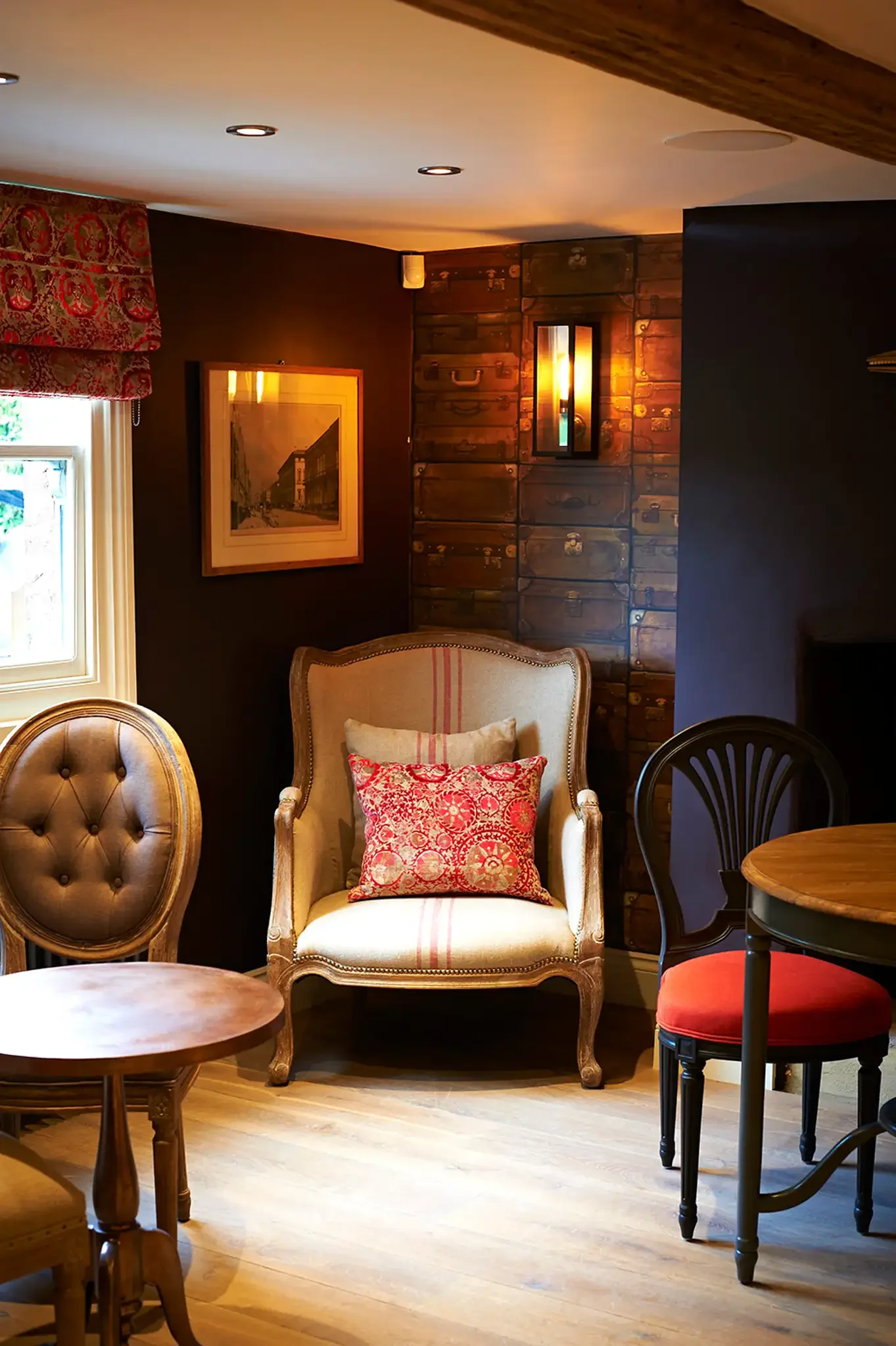 Interior of a cozy living room with wooden floors, vintage furniture including a tufted armchair and a striped armchair with cushions, a wooden dining table, and wall decor such as framed art and a decorative wall light, with a window on the left letting in natural light.