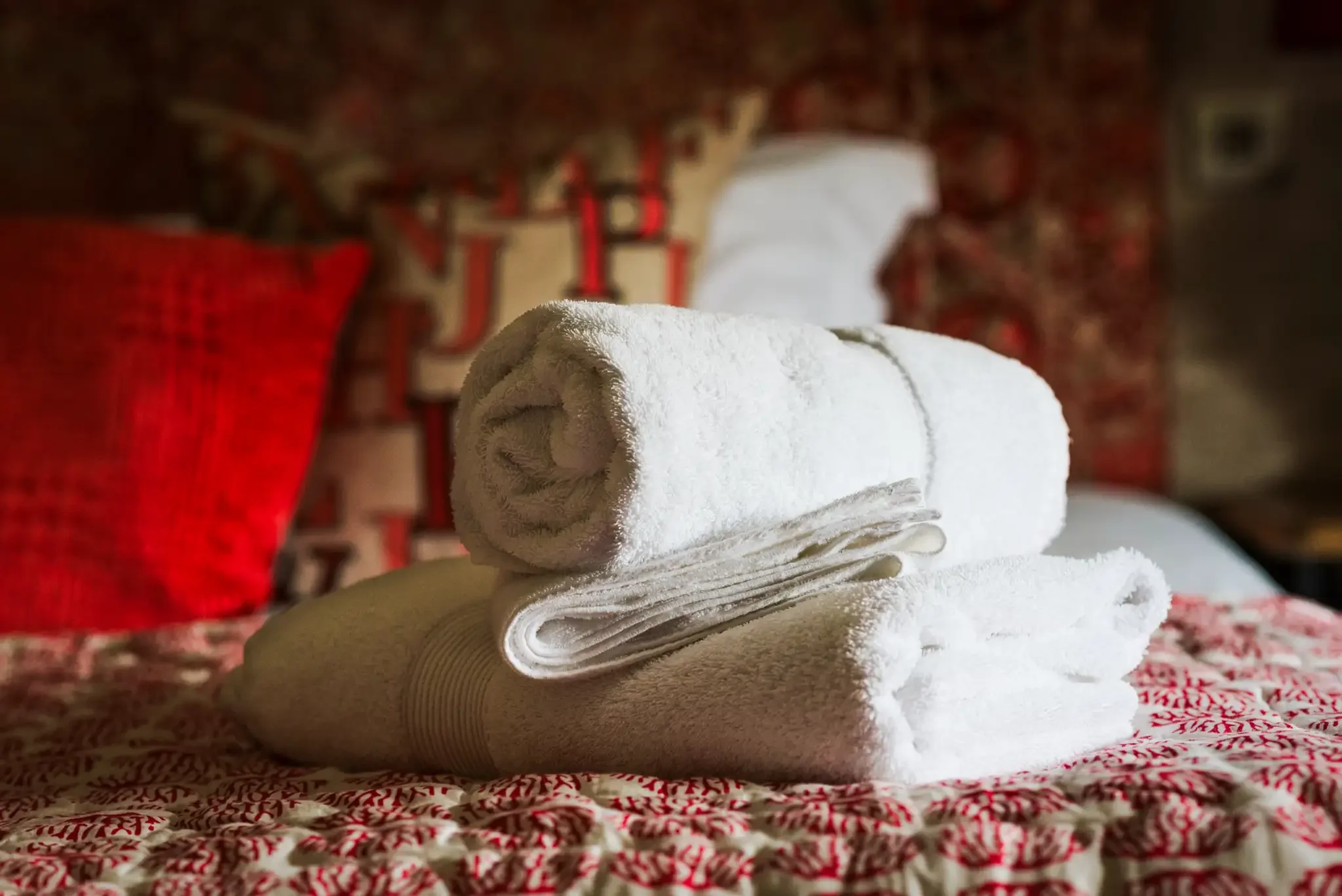 Stacked white towels on a patterned bedspread with red pillows and a decorative headboard in the background.