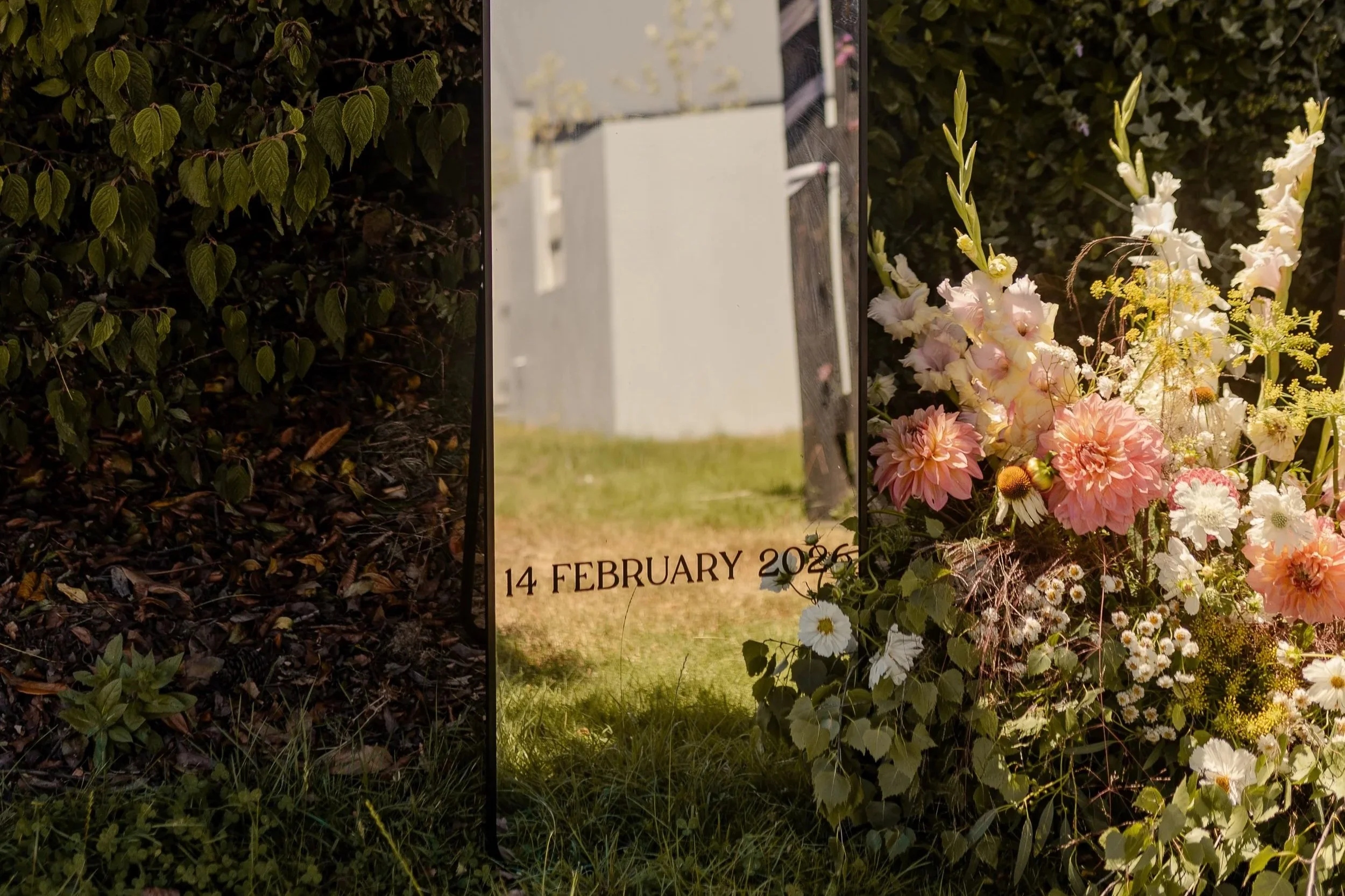Photograph of floral arrangements with pink and white flowers alongside greenery, and a memorial sign with the date '14 February 2026.'