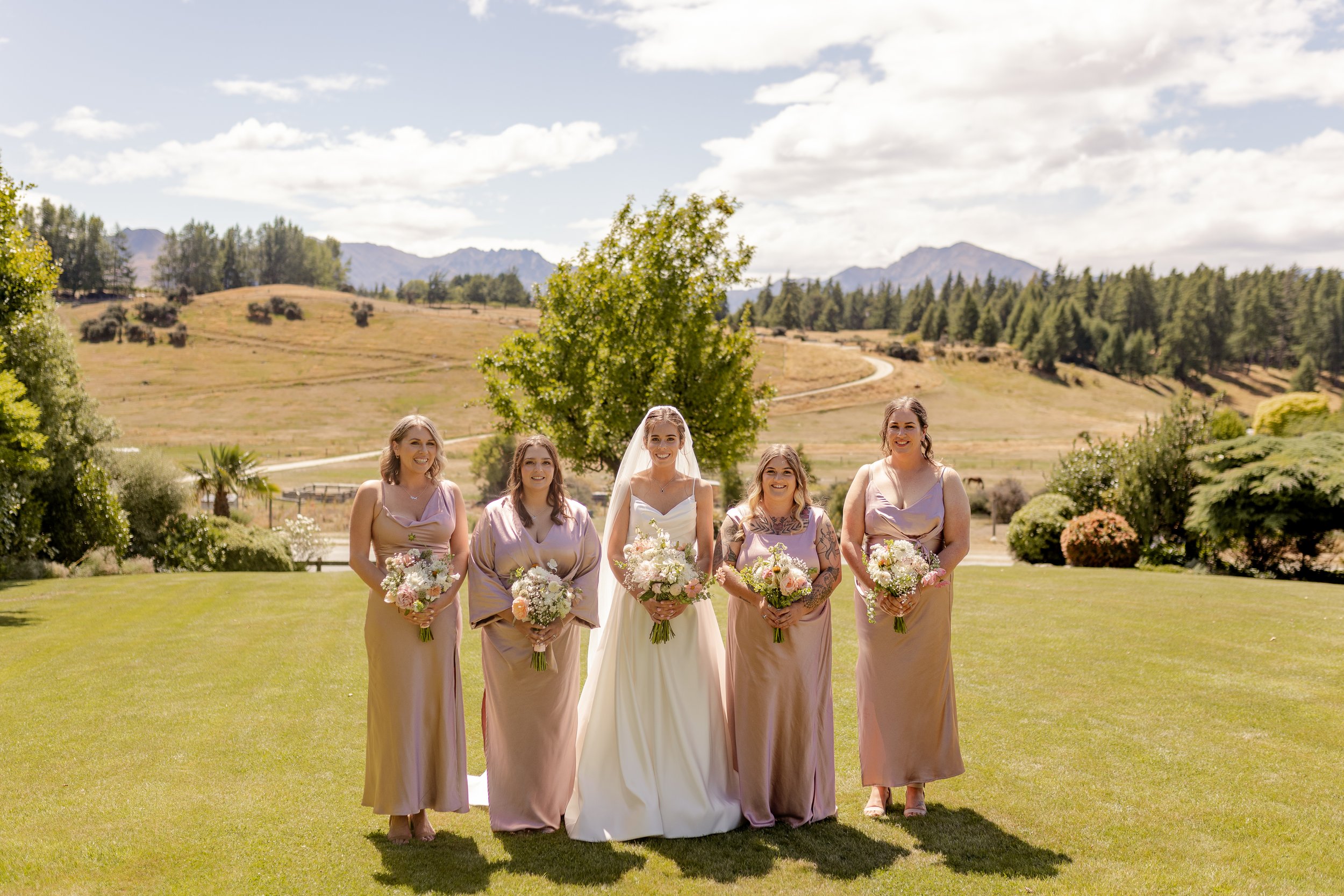 A group of women, including a bride in a white dress and veil, and four bridesmaids in mauve dresses, standing on a green lawn outdoors with mountains and trees in the background, during a wedding celebration.