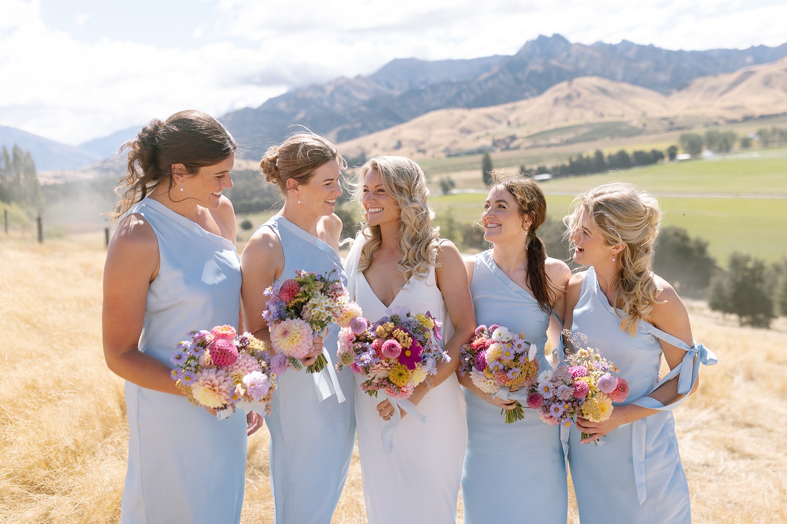 A group of five women in bridesmaid and bride dresses, holding bouquets, standing in a field with mountains in the background, smiling and interacting with each other.