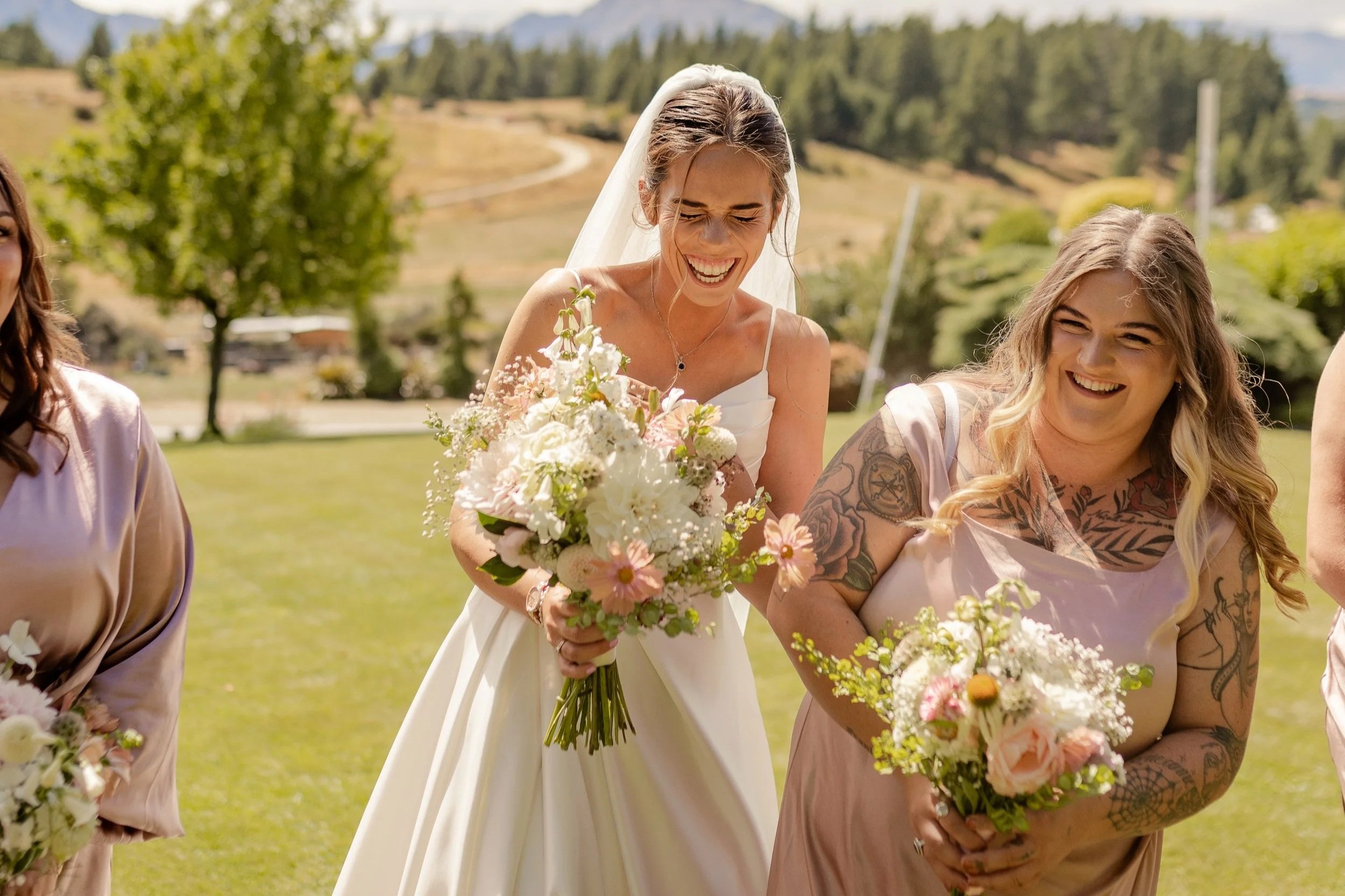 A bride in a white dress holding a bouquet of flowers, smiling, surrounded by women holding bouquets, outdoors on a sunny day with trees and rolling hills in the background.