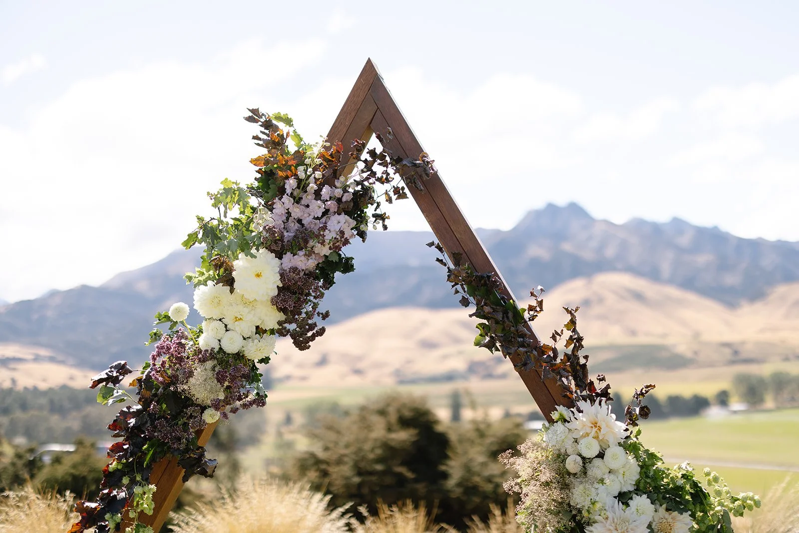 A wooden wedding arch decorated with white and purple flowers, set outdoors against a mountain landscape and a partly cloudy sky.