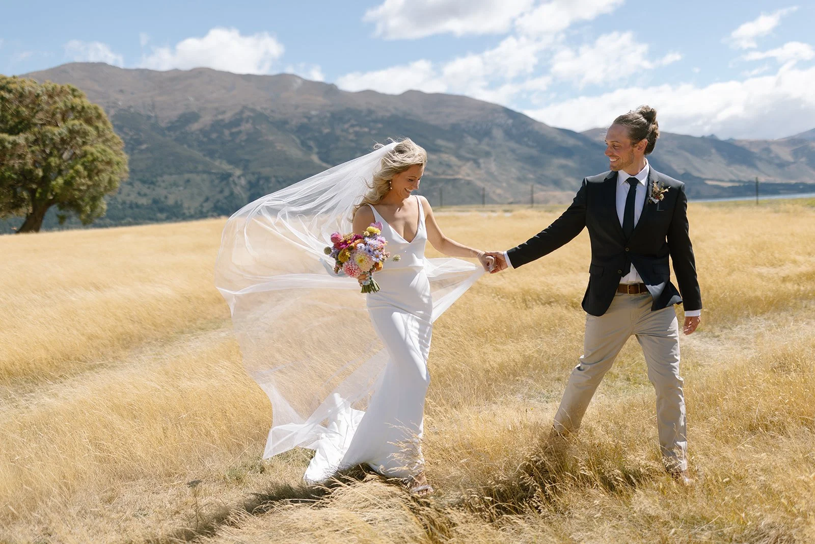 A bride and groom holding hands and dancing in a field with mountains in the background.