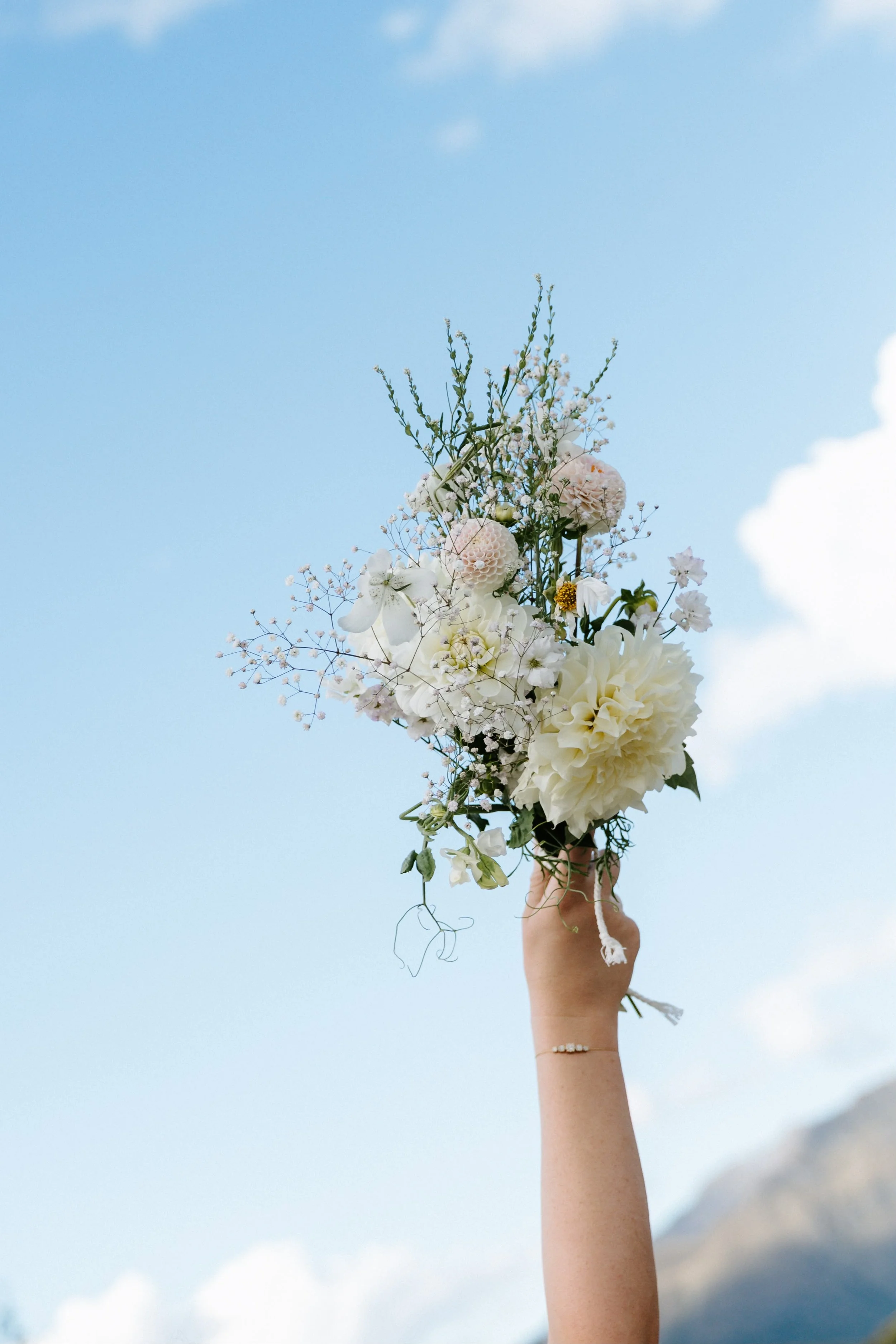 A hand holding a bouquet of white and pink flowers against a blue sky with clouds.