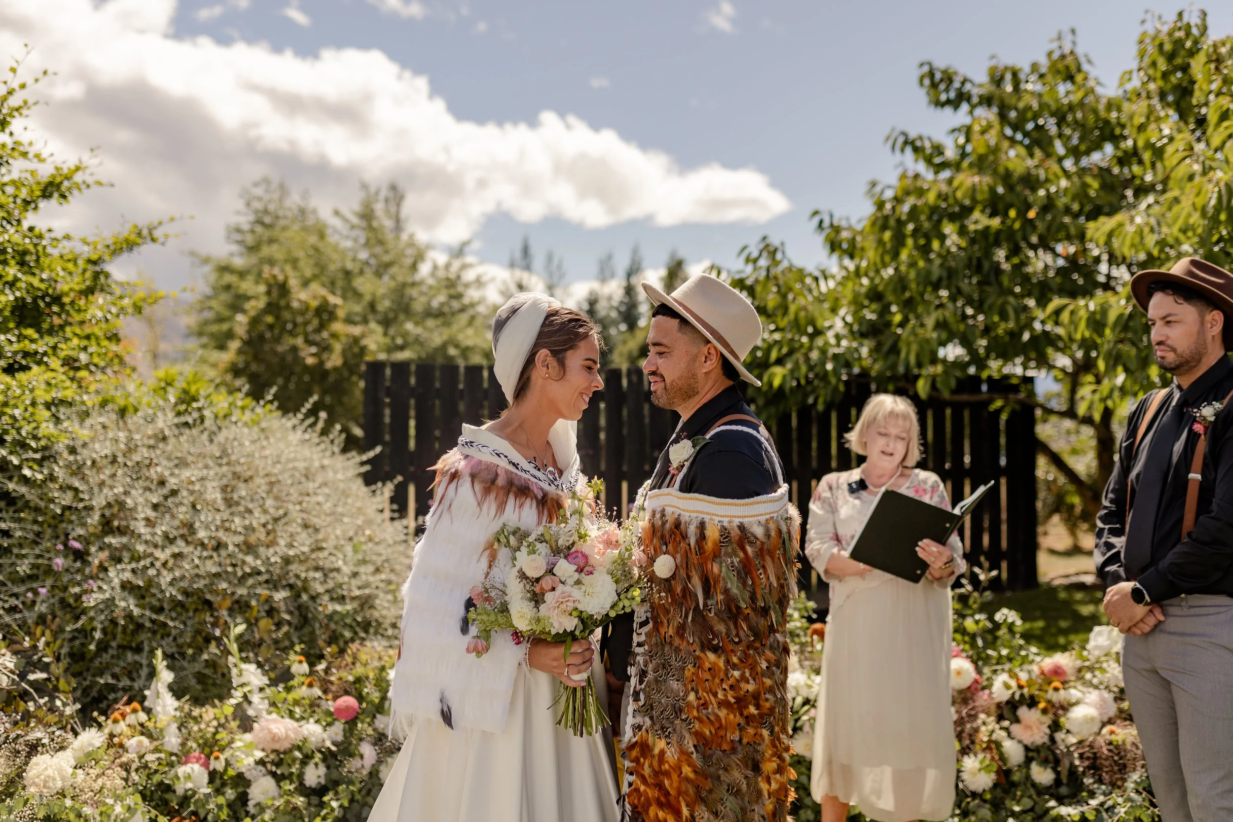 A couple exchanging wedding vows outdoors surrounded by flowers, with guests and officiant in the background.