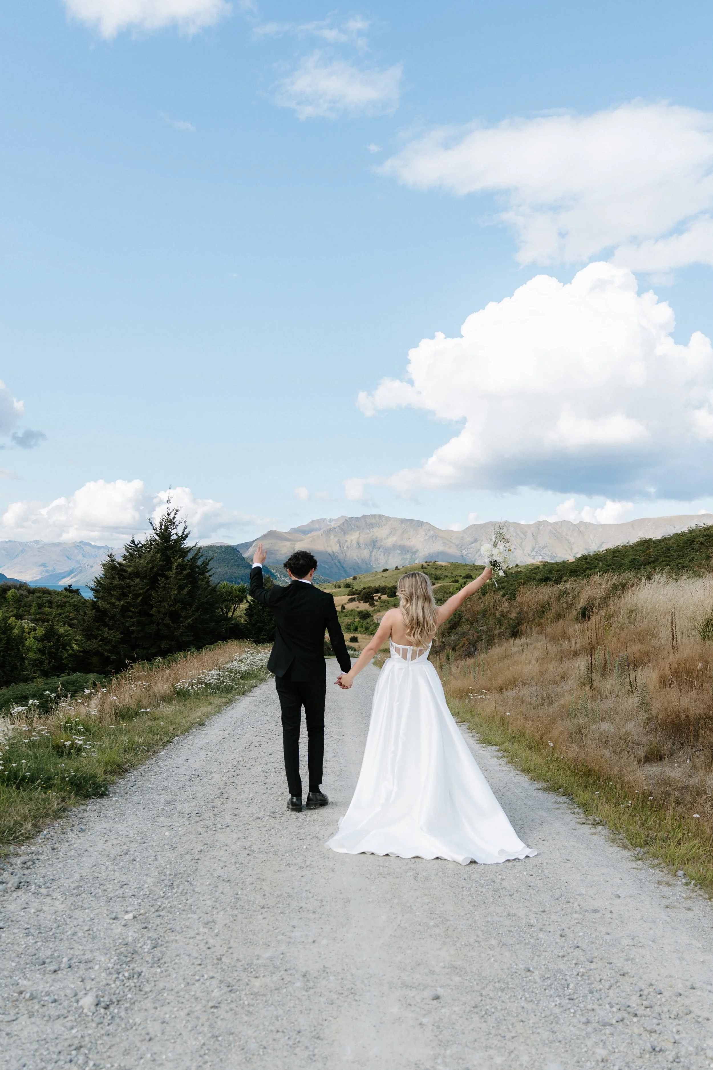 A newlywed couple, the groom in a black suit and the bride in a white wedding dress, holding hands and walking away on a gravel dirt road in a rural landscape with mountains and hills, under a partly cloudy sky.