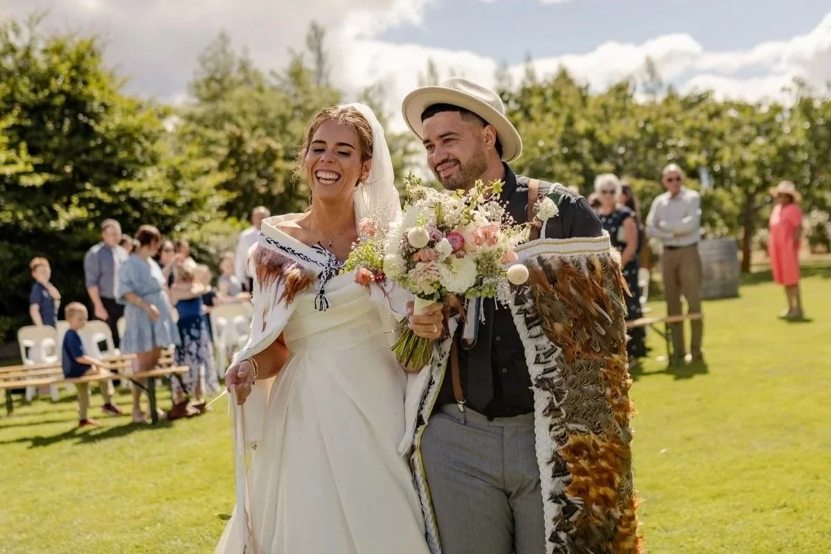 A newlywed couple, the bride in a white wedding dress and the groom in a dark shirt with a straw hat, walking arm in arm outside at wedding or celebration, holding a bouquet of flowers, smiling, with guests seated and standing in the background on a sunny day.